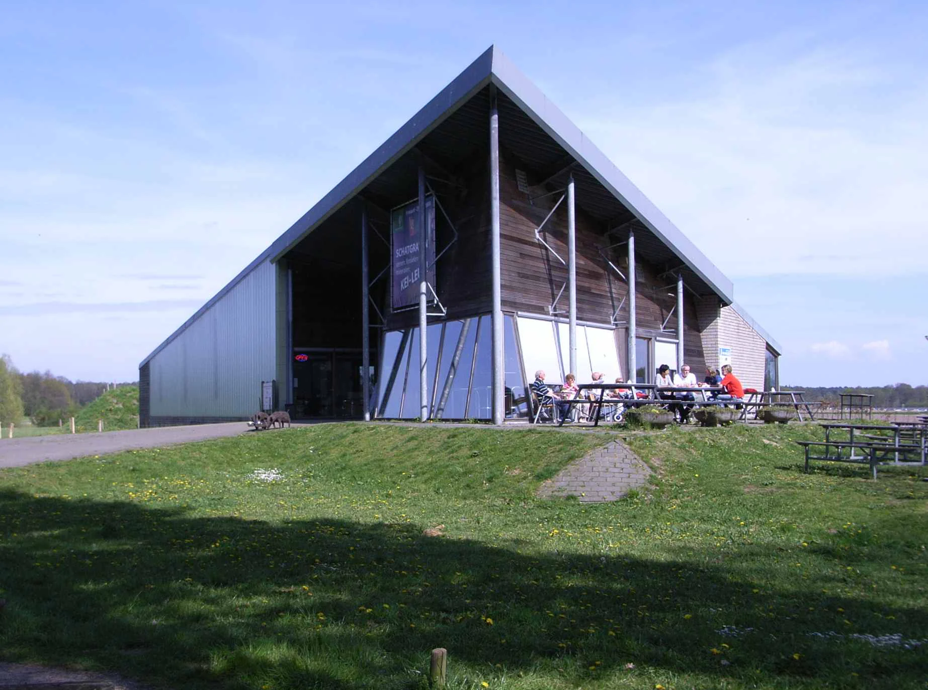 A modern visitor center building with a large triangular roof and glass facade situated in a grassy park setting, with people sitting at outdoor picnic tables.