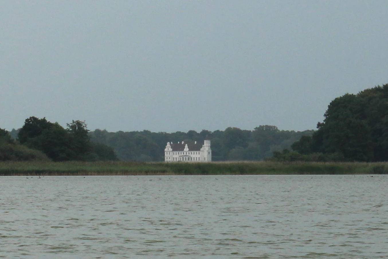A white manor house with a classic gabled roof sits on the distant shore of a large lake, surrounded by dark green trees under an overcast sky.