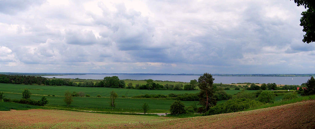Panoramic landscape view of a large lake in the distance, framed by green meadows, cultivated fields, and trees under a cloudy sky.