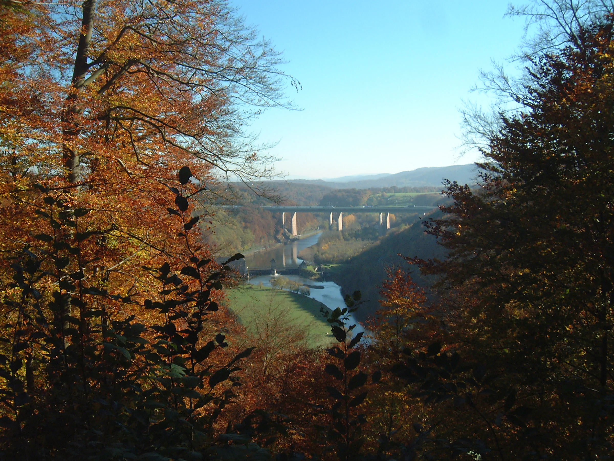 A valley view through an autumn forest featuring a river and long viaduct bridges in the distance.
