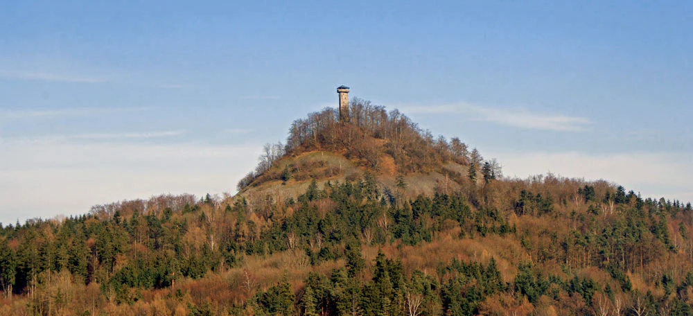 Una vista lejana de una montaña cónica, el Rauher Kulm, con una torre de observación alta en su cima, rodeada de un paisaje boscoso.