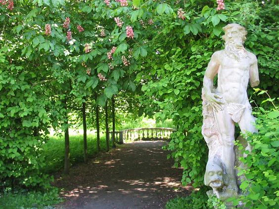 Un camino de grava bordeado por castaños de Indias verdes con grupos de flores rosadas conduce hacia una valla de madera a lo lejos, mientras que una estatua de piedra blanca desgastada que representa una figura masculina se alza entre la vegetación frondosa en el lado derecho.