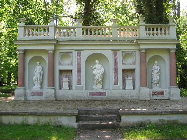 Un muro de piedra de estilo clásico con tres nichos arqueados que contienen estatuas blancas, columnas rosadas y una balaustrada superior, situado en un parque verde con árboles al fondo y escalones que conducen a él.
