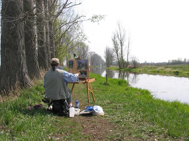 Una persona sentada en un taburete pintando sobre un caballete a la orilla de un río, con una hilera de grandes árboles a la izquierda.