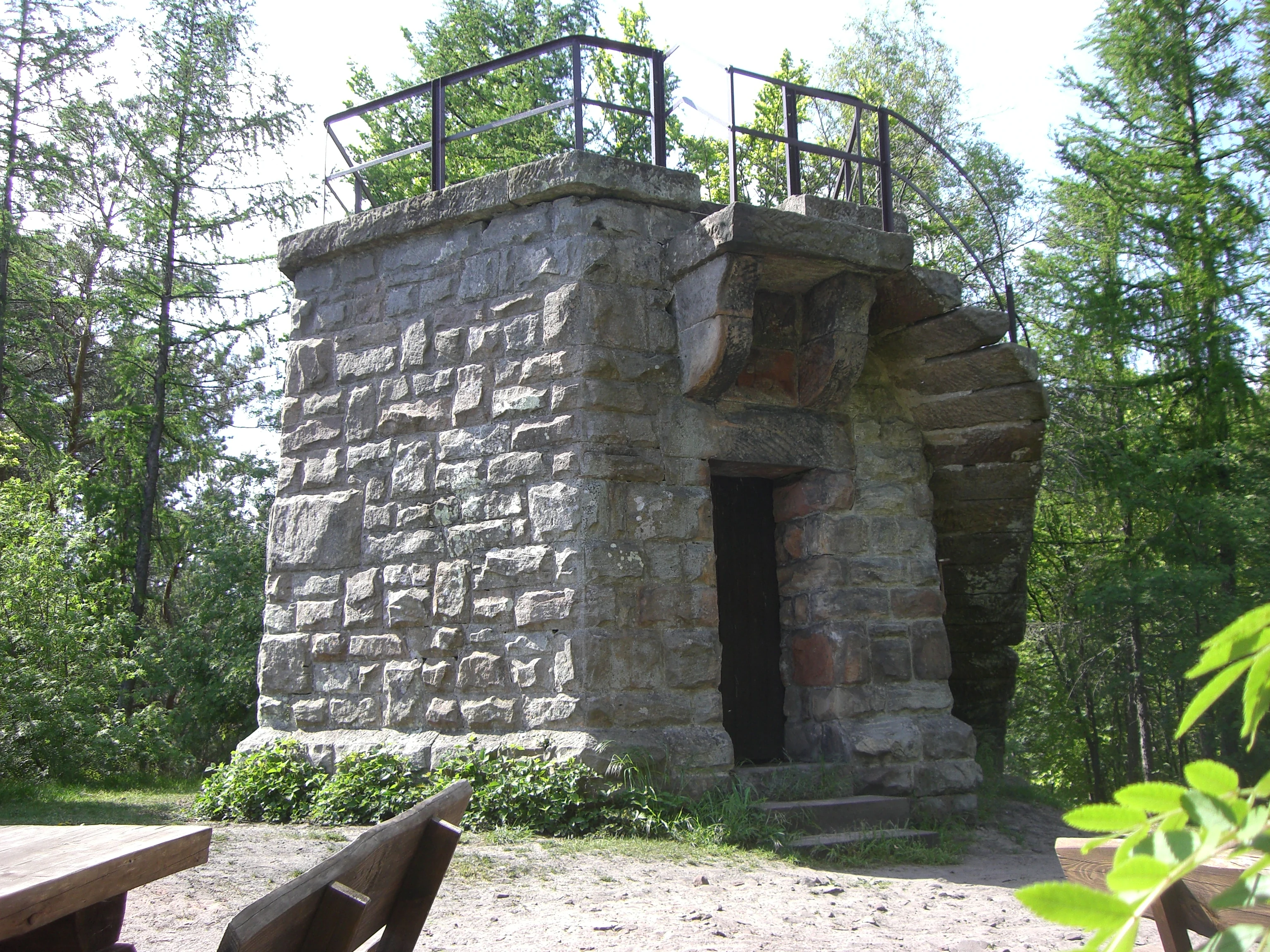 A stone observation tower with a square base and open-air viewing platform, surrounded by forest trees.