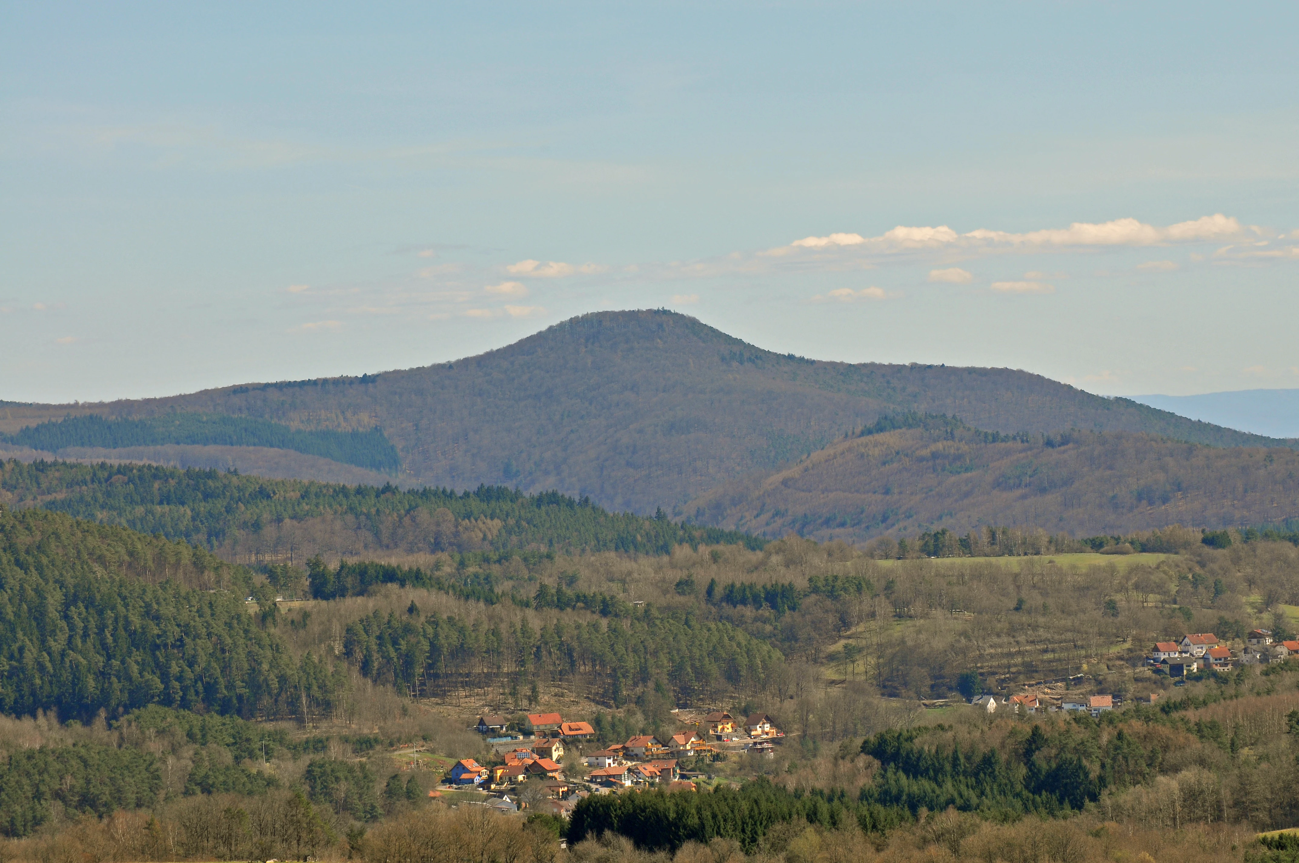 A wide mountain landscape showing a rolling forested hill in the center, a small town with red-roofed buildings in the valley, and distant hills under a clear blue sky.