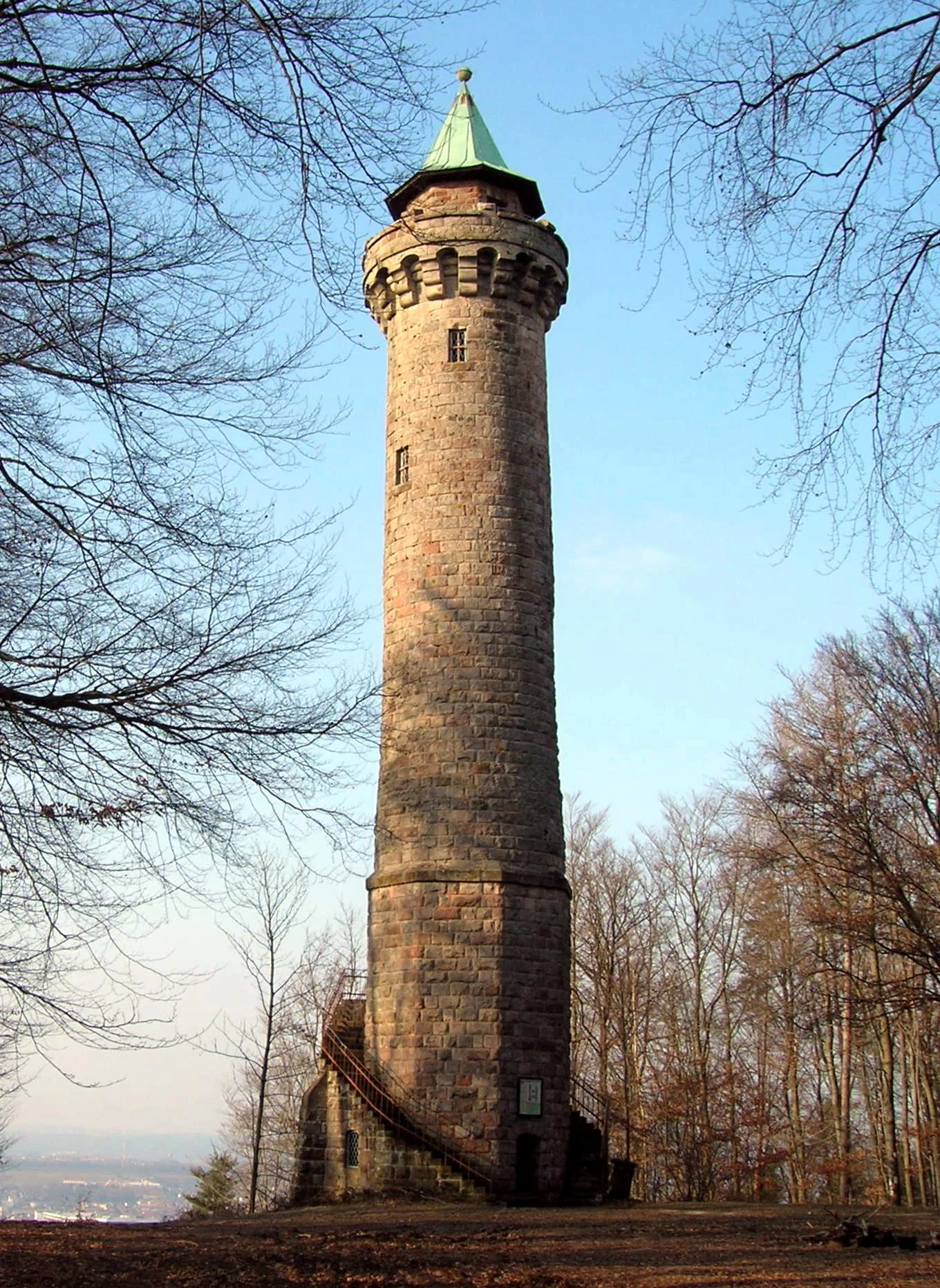 A tall stone observation tower with a green pointed roof stands in a forest, framed by leafless tree branches against a clear blue sky.
