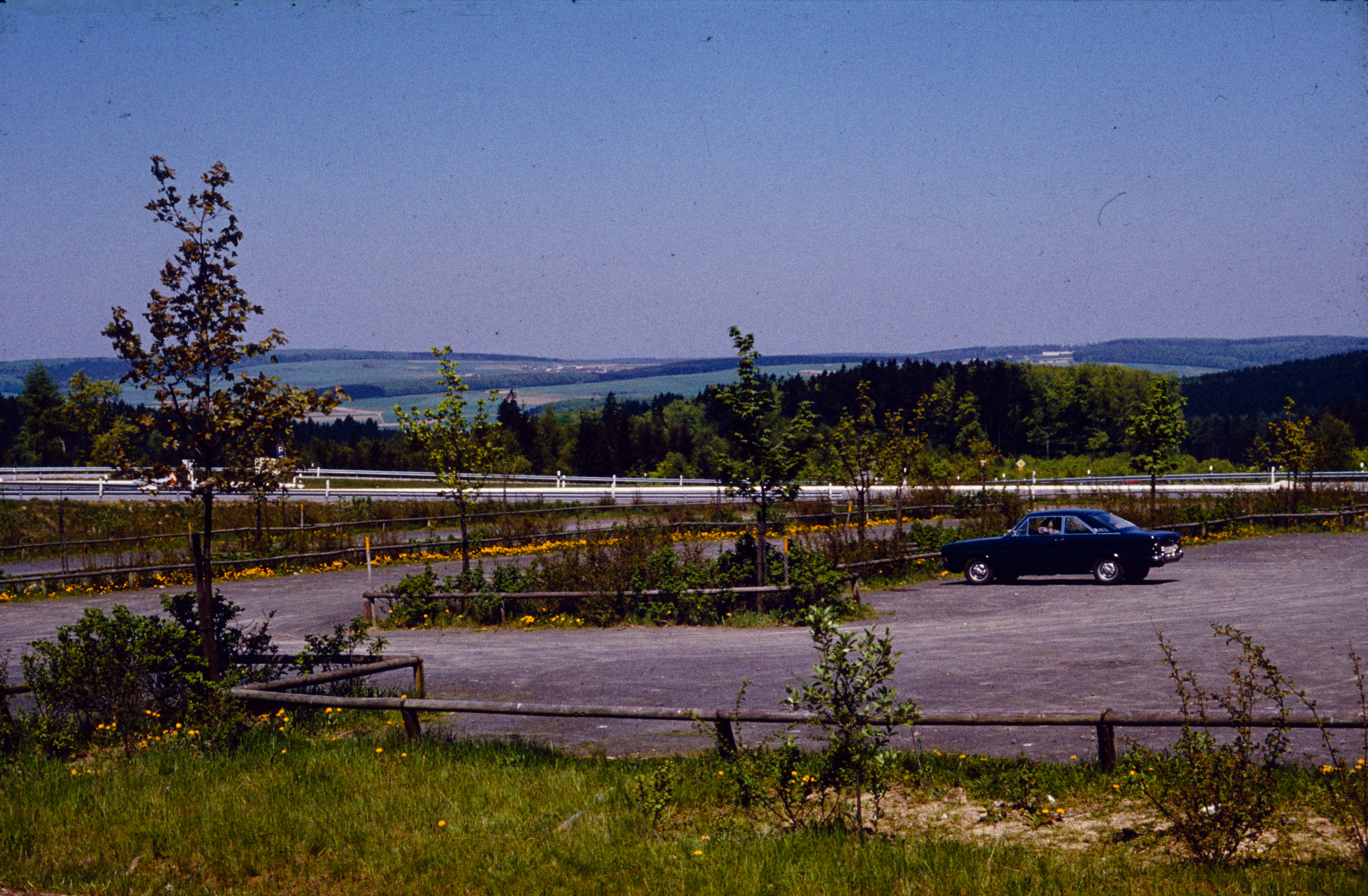 A vintage color photograph showing a gravel parking area with a lone dark car, surrounded by young trees and wooden fencing, overlooking a distant hilly forest landscape under a clear blue sky.