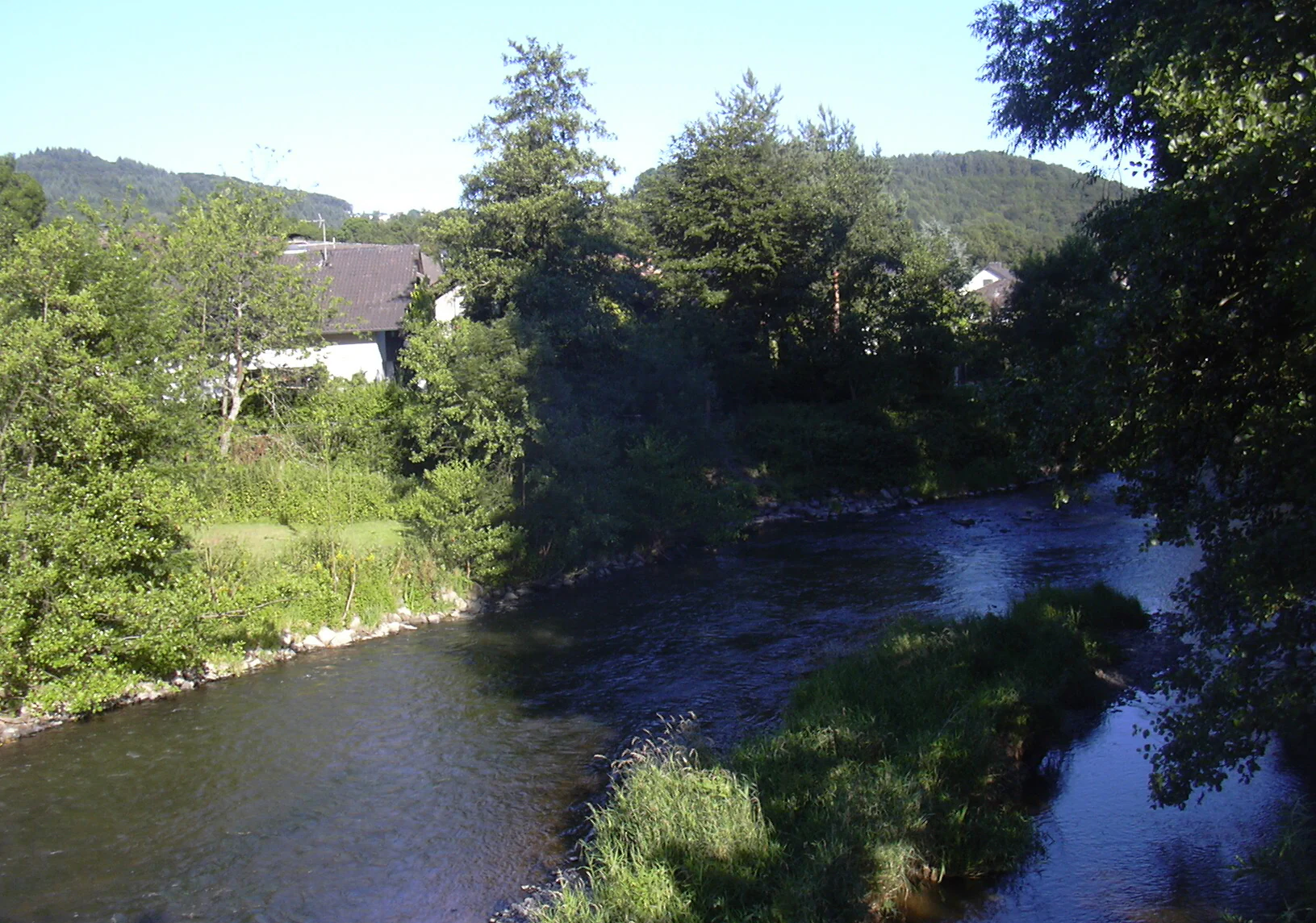 A view of the Wied river flowing through a forested landscape with a small island of vegetation in the foreground and a house visible in the trees.