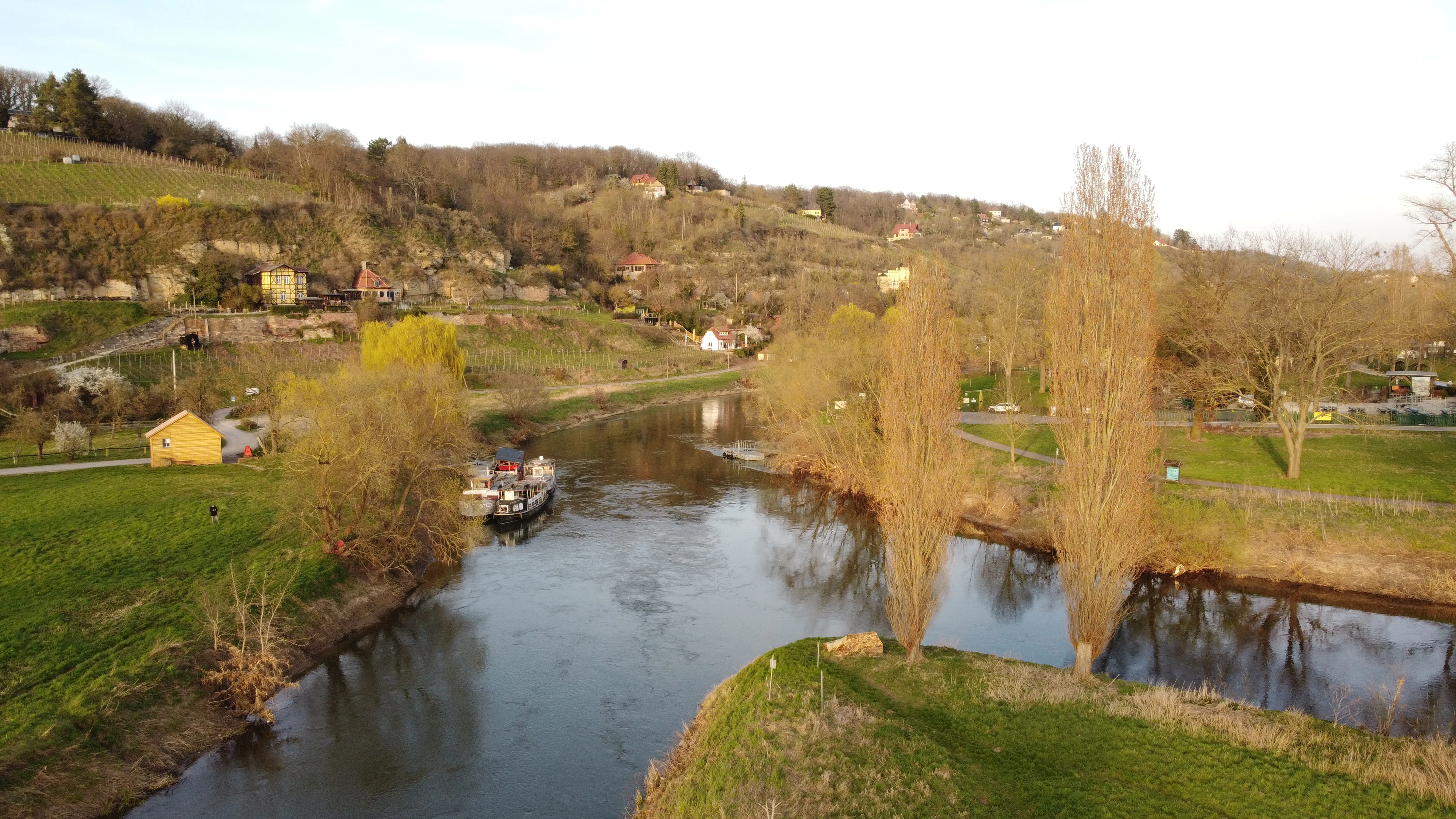 An aerial view of the confluence of two rivers in Germany, showing green banks, tall thin trees, a small boat docked, and hillsides in the background.