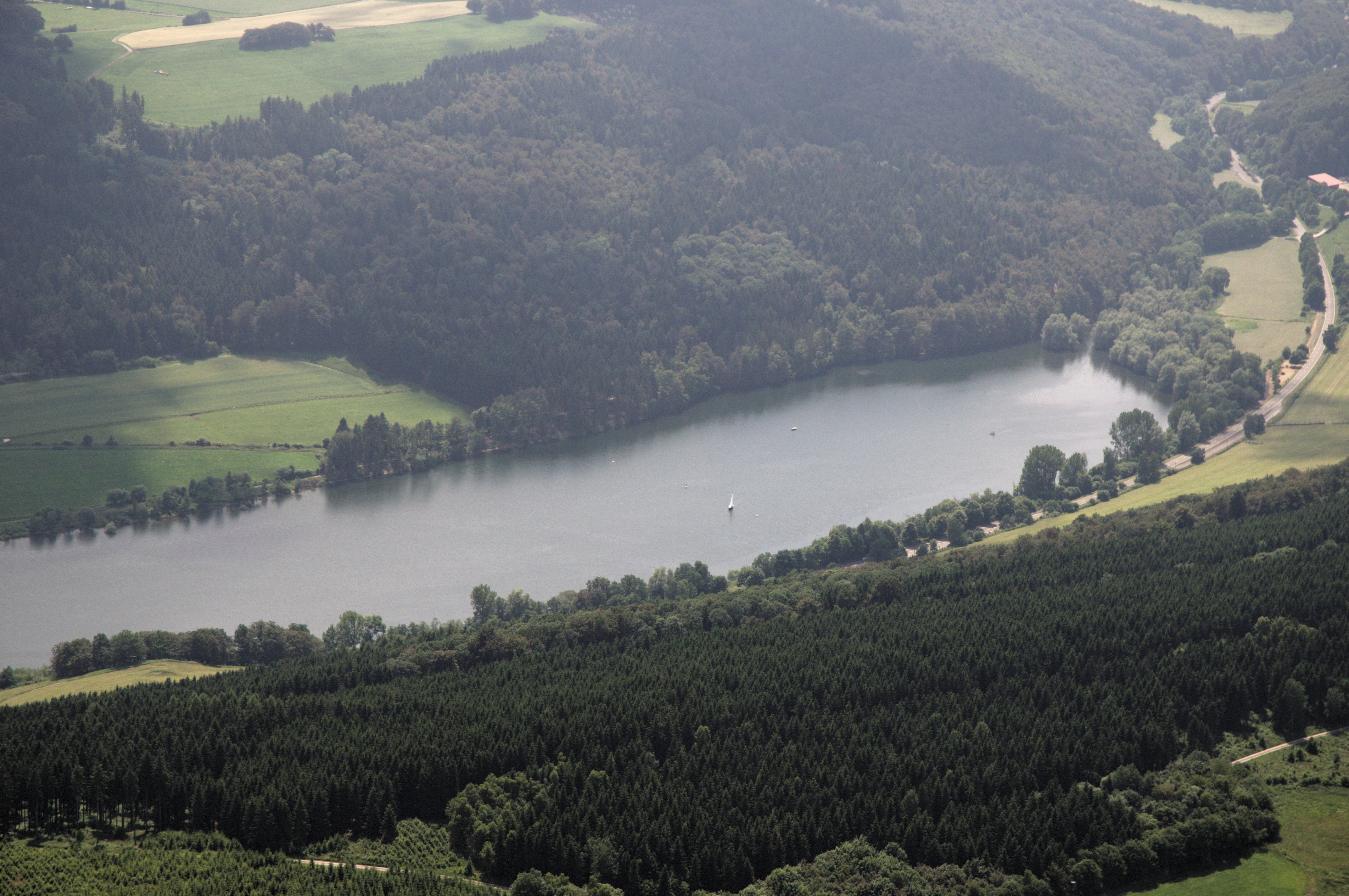 Aerial perspective of a calm lake surrounded by dense forests, meadows, and a winding road in the Sauerland countryside.
