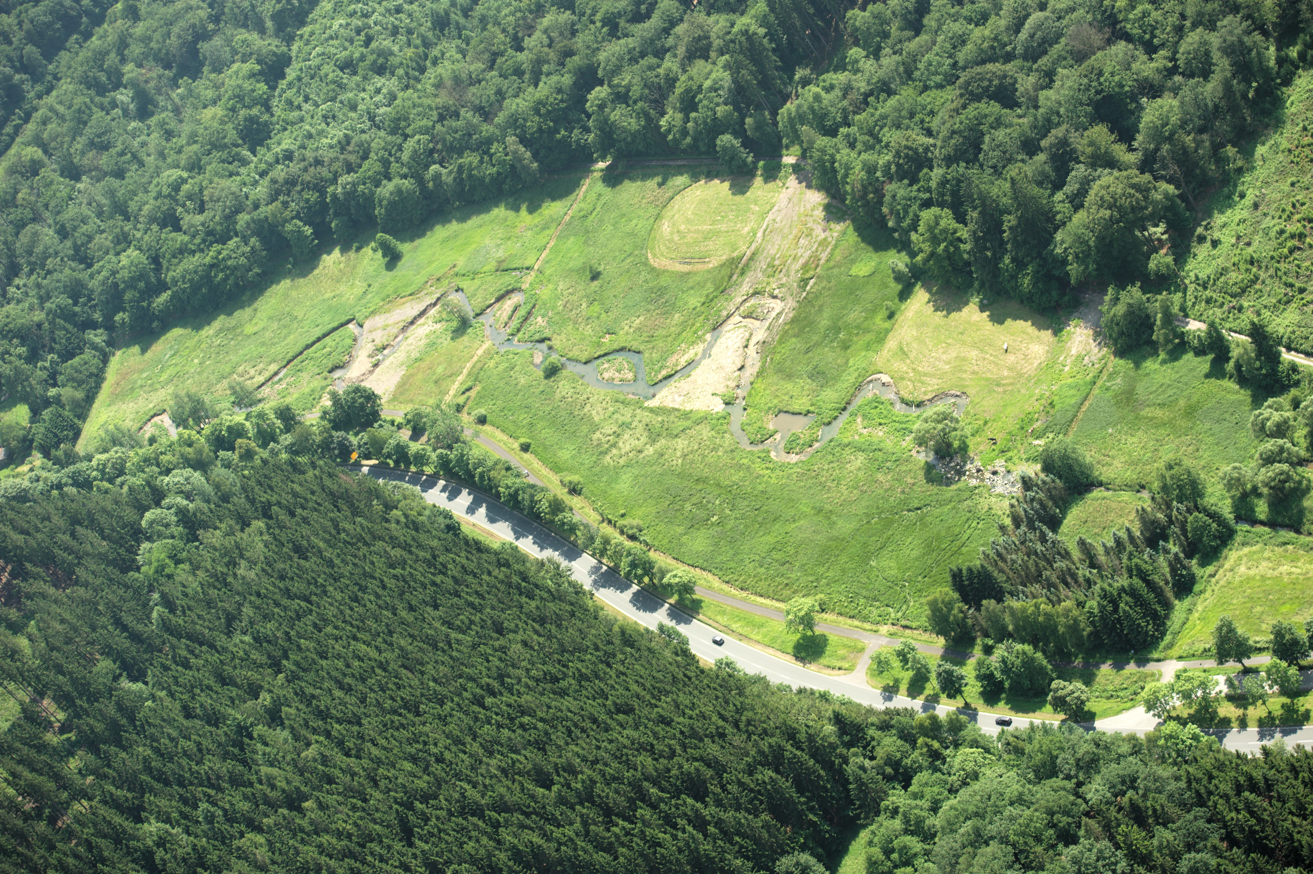An aerial shot showing a winding river segment in a green valley, surrounded by dense forest, with a road running along the lower edge.