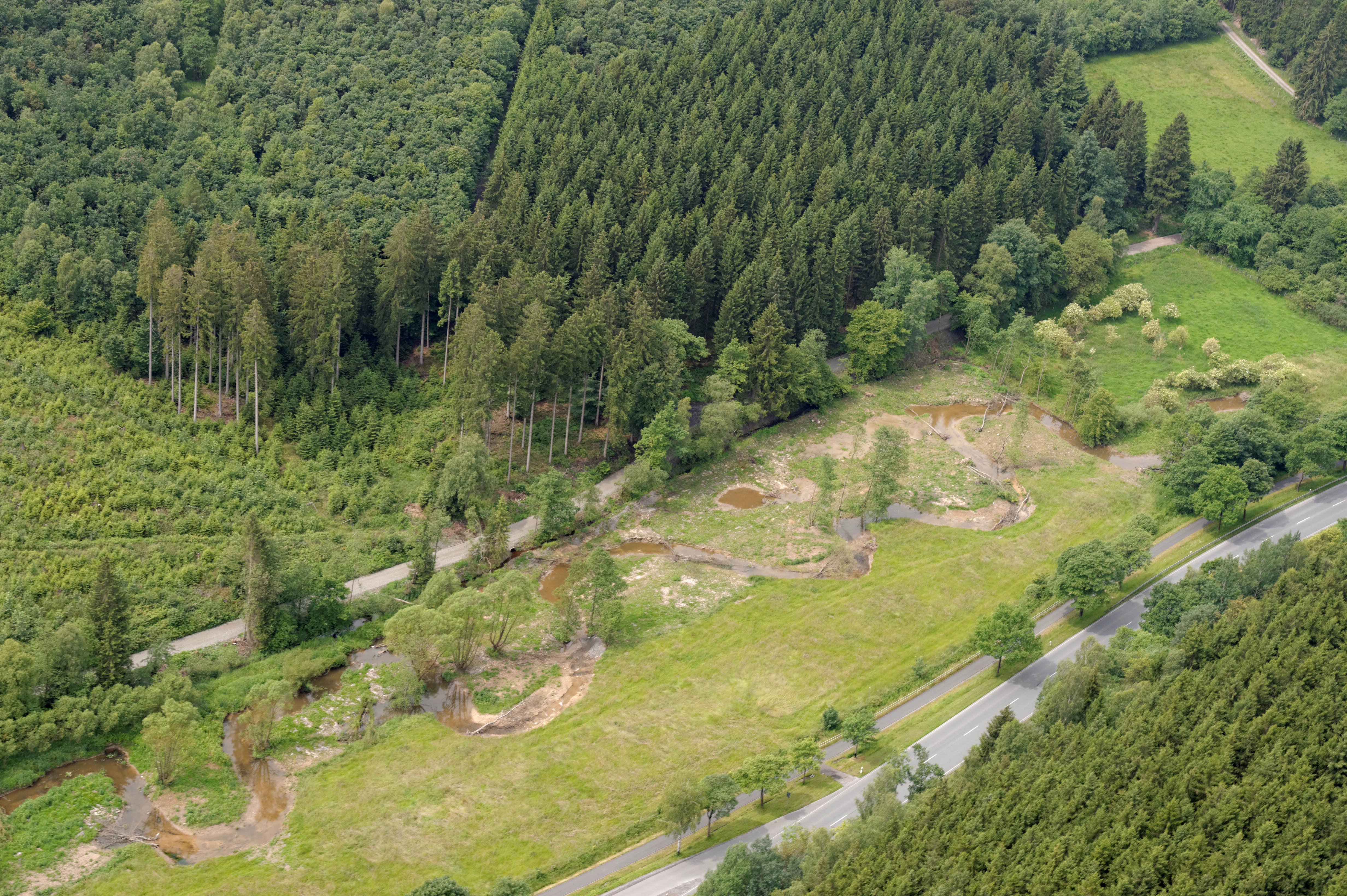 Aerial view of a narrow river flowing through a green river valley, with forests on both sides and a road running parallel to the water.