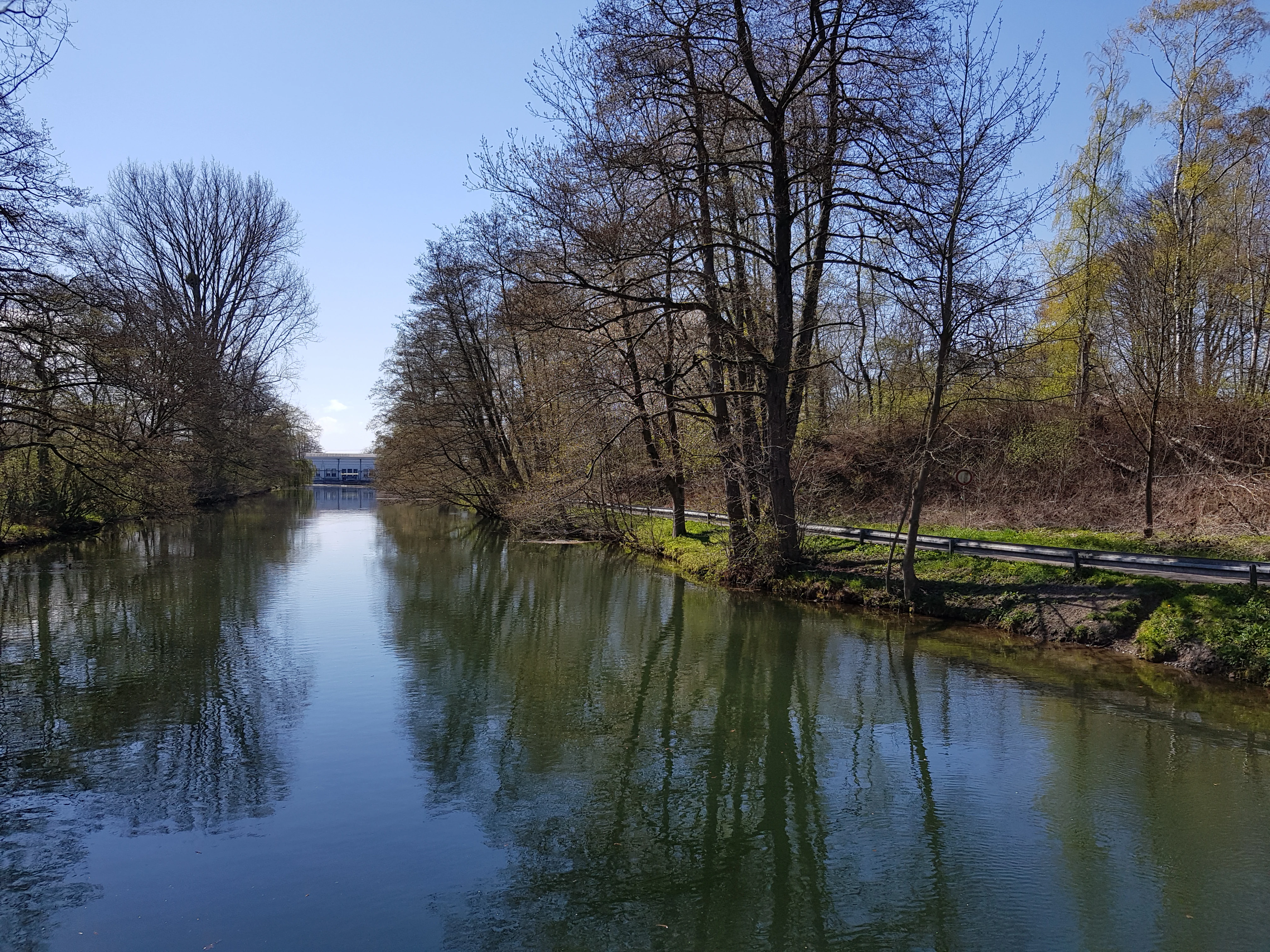 A calm, narrow canal or river flowing through a lush wooded area with reflections on the water surface and a building visible in the distance.