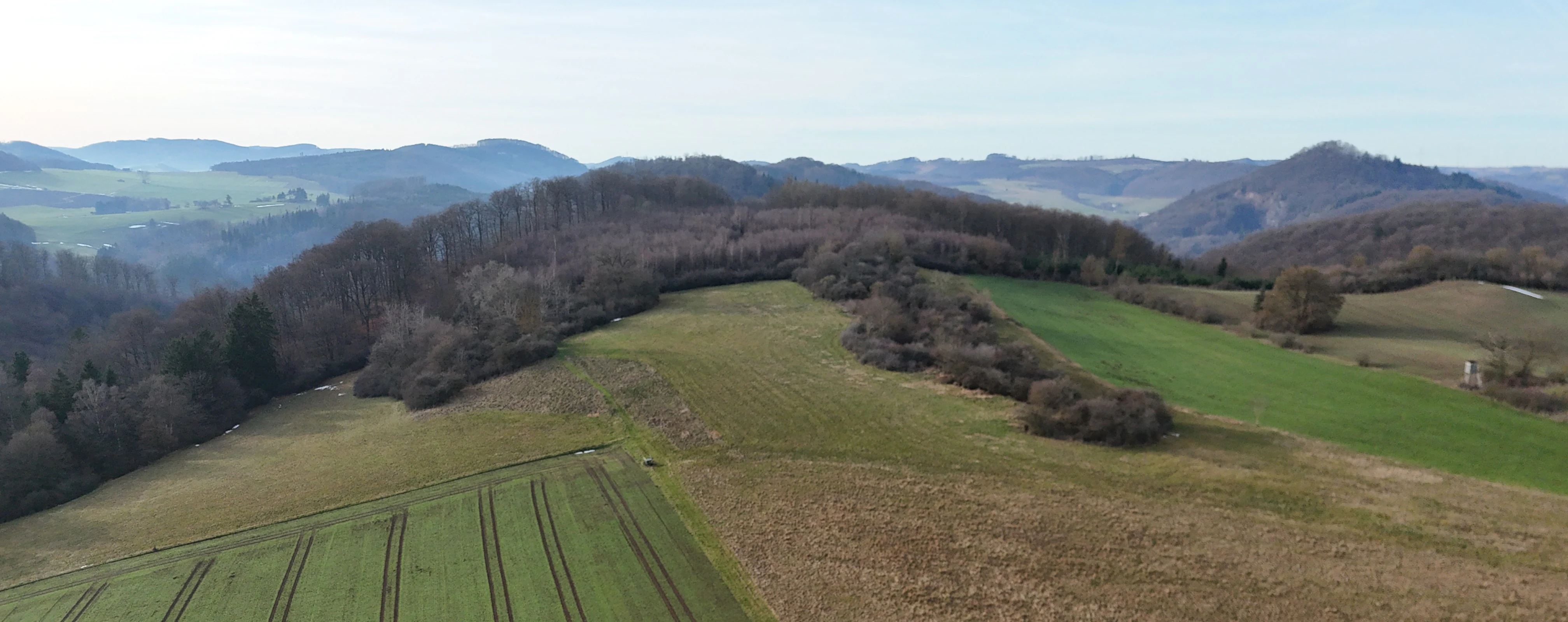 A wide aerial shot of a hilly landscape featuring green agricultural fields in the foreground, transitioning into patches of scrub and deciduous forest, with soft blue hills visible on the horizon.