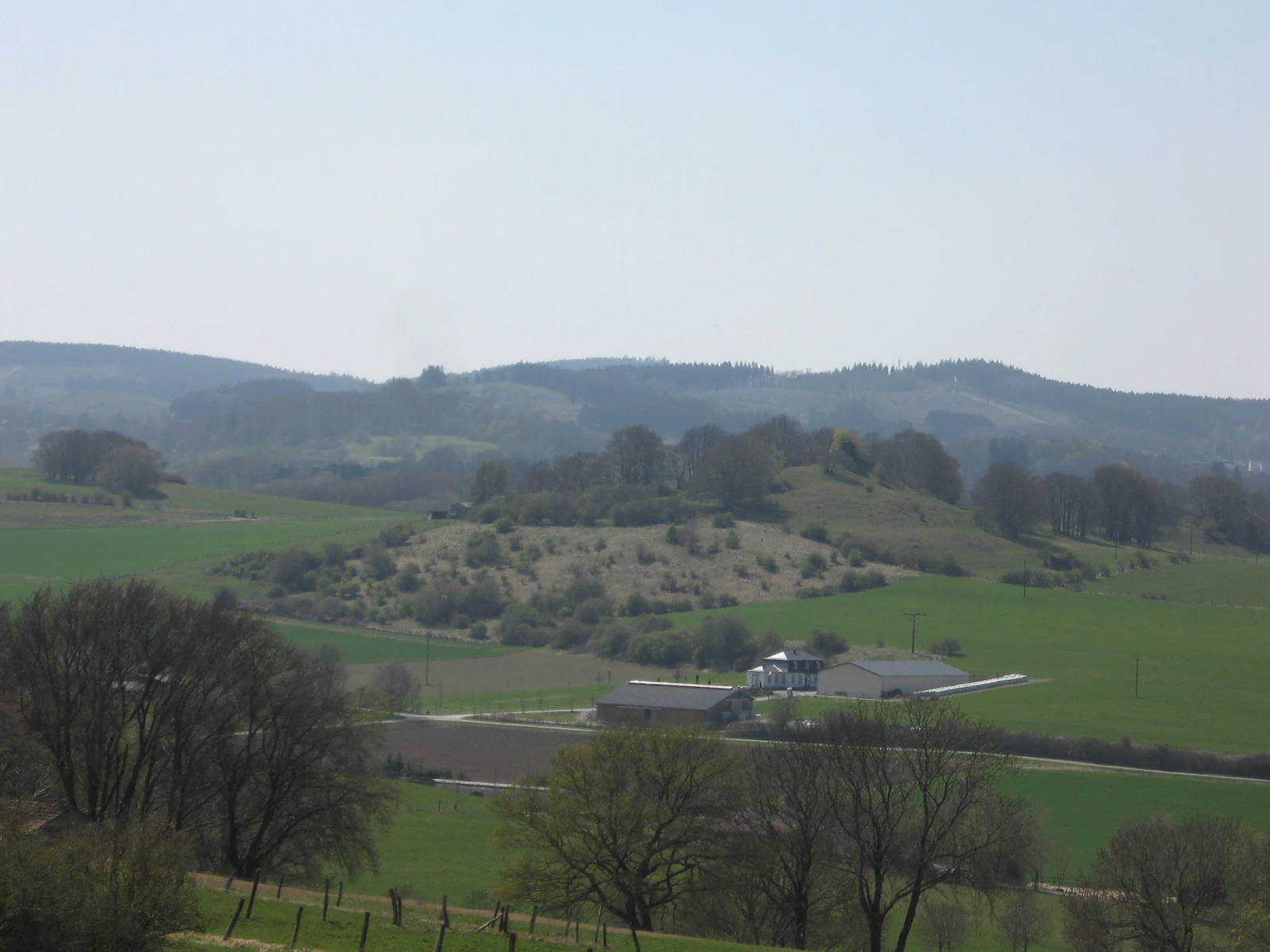 A wide landscape view of rolling green hills and fields with scattered trees and a farm complex in the middle distance under a hazy sky.