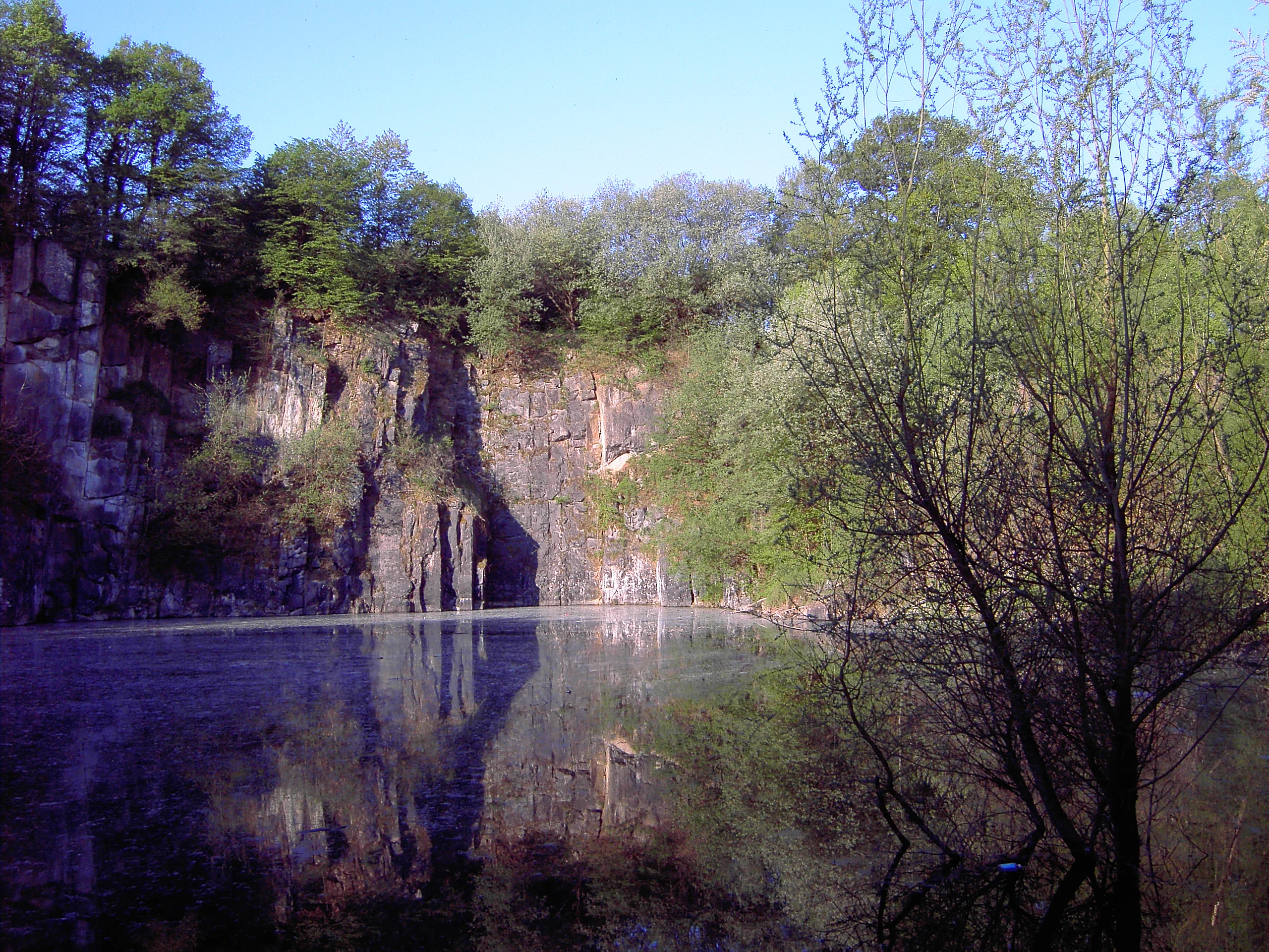 A lake with still water reflecting rocky quarry cliffs and surrounding trees under a clear blue sky.