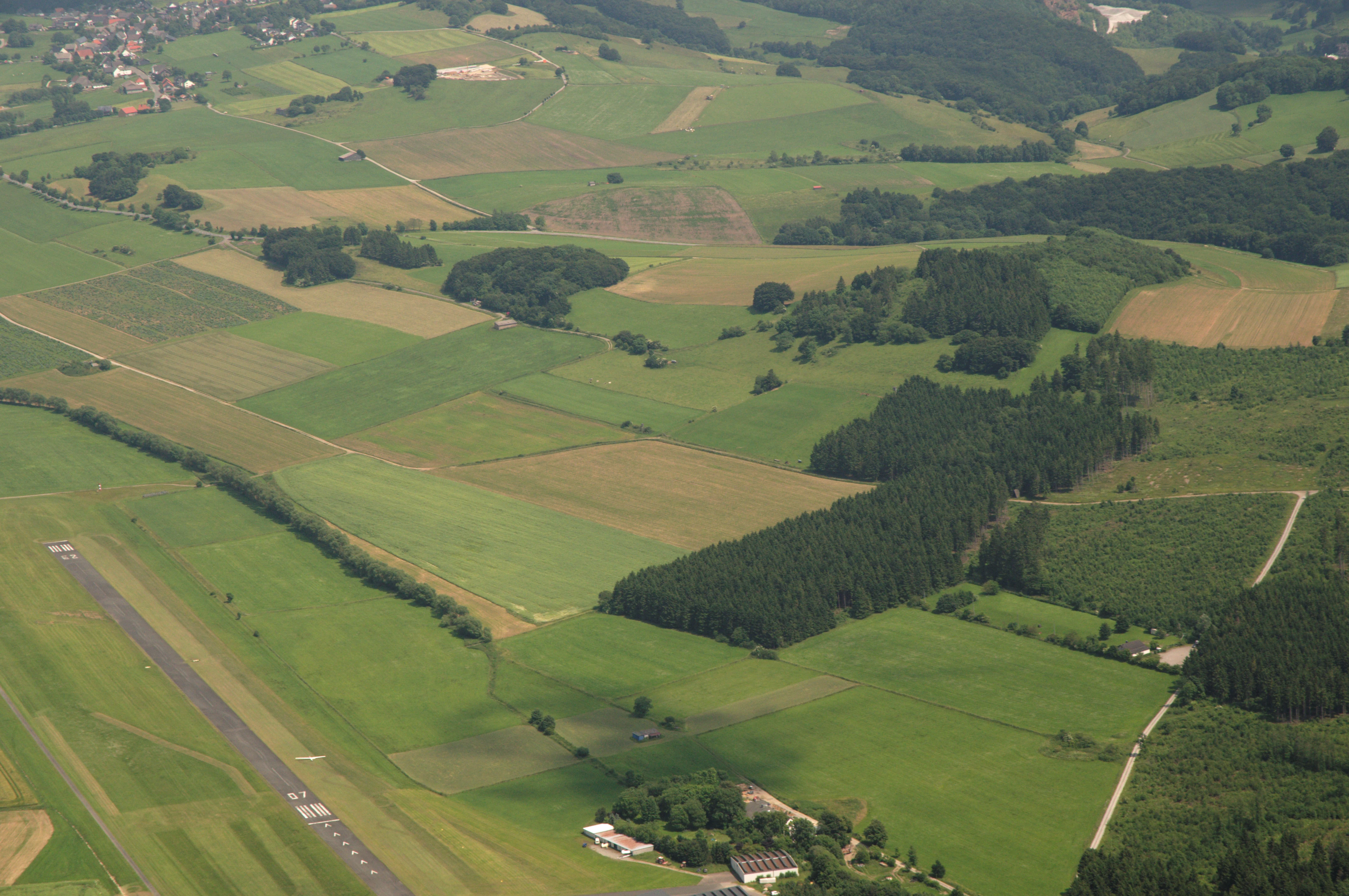 Fotografía aérea en picado que muestra un mosaico de campos agrícolas verdes, pequeñas zonas boscosas y una pista de aterrizaje pavimentada en la esquina inferior izquierda.