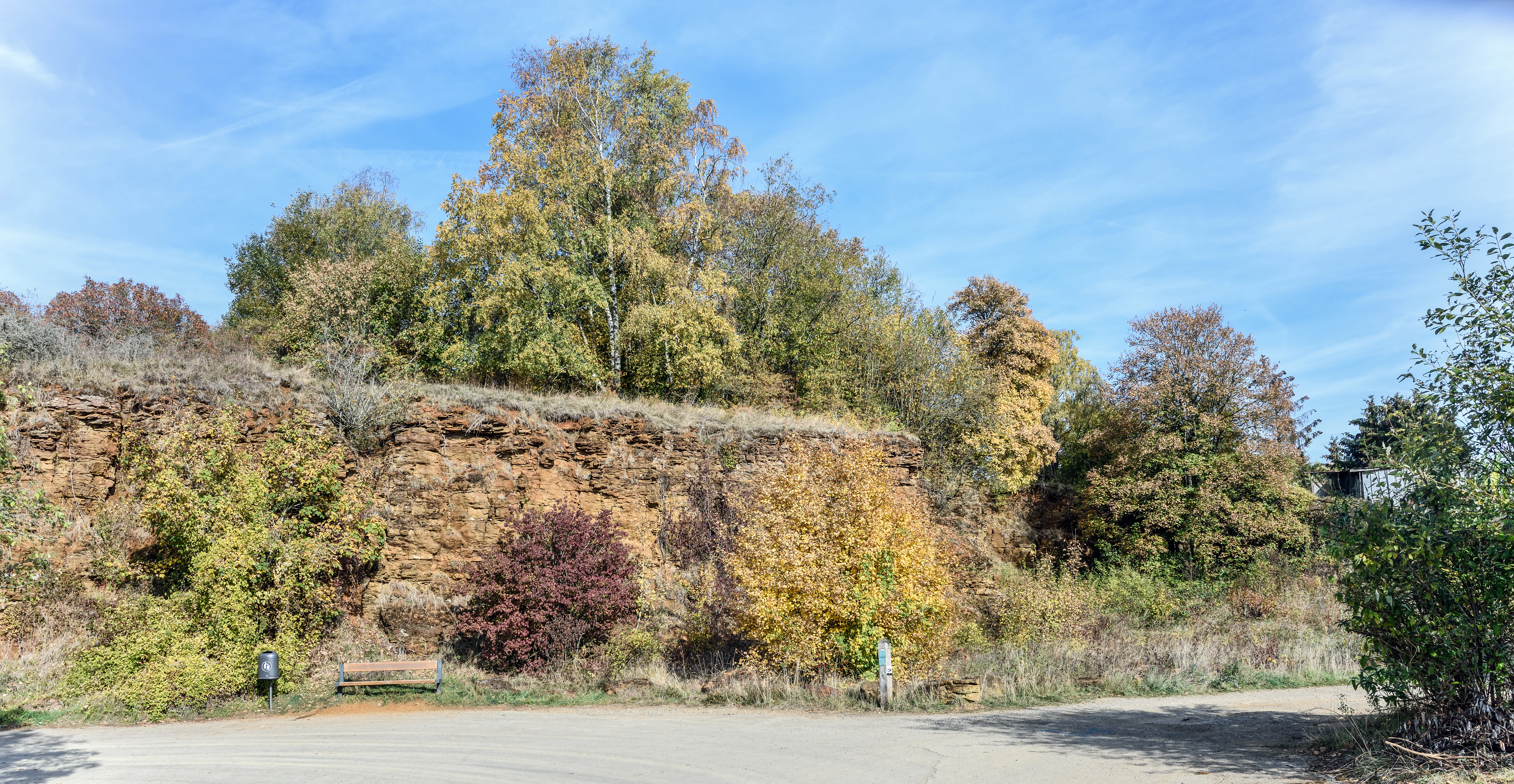 A dirt path curves toward a low, rocky cliff face covered in autumn-colored trees and brush.