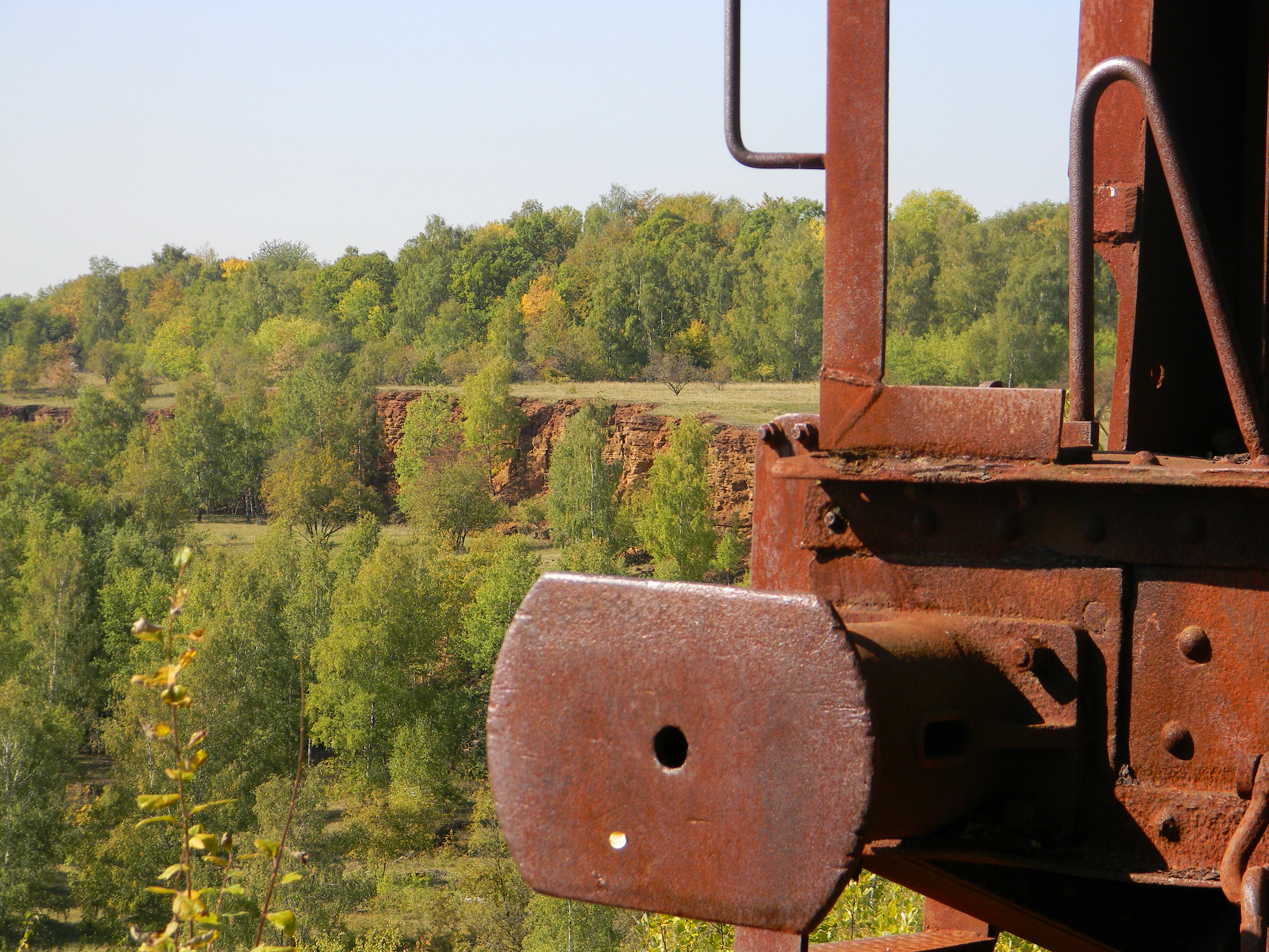 A close-up view of a rusty, weathered metal mining machine component in the foreground, with a background of trees and a rocky cliff face in a nature reserve.