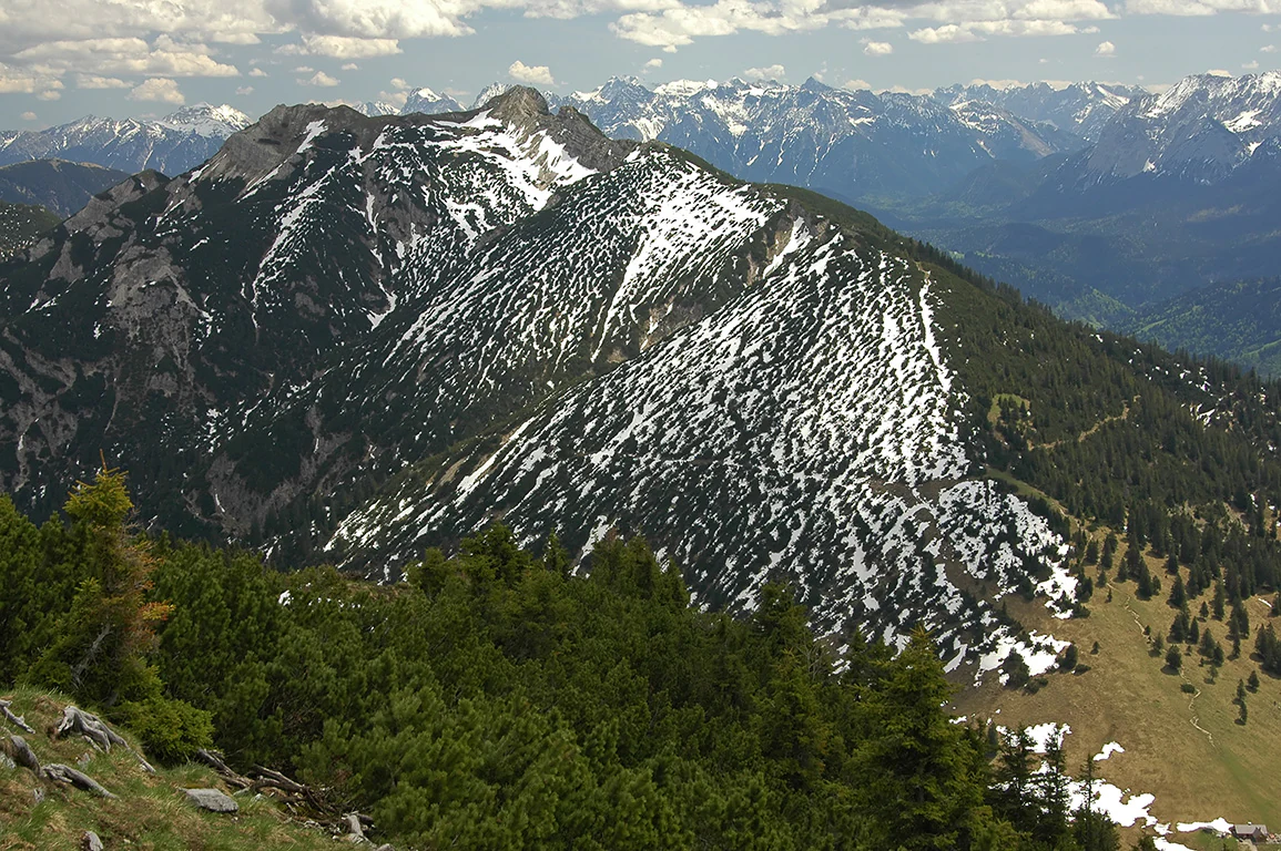 High altitude view of the rocky, snow-dusted Kramerspitz peak and surrounding forest in the Ammergau Alps.