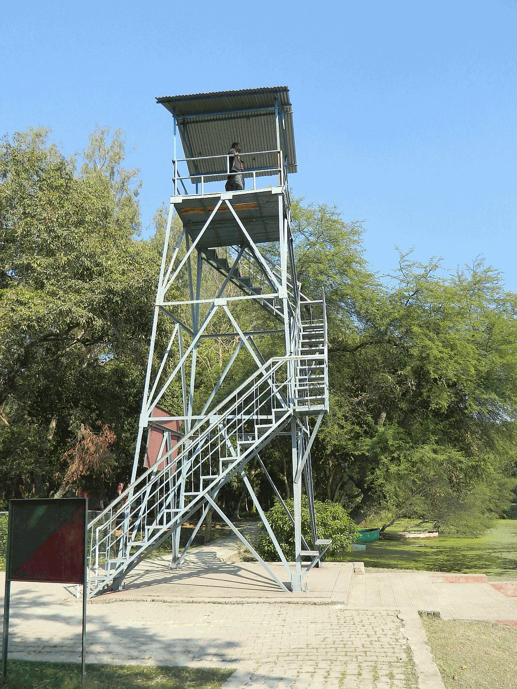 A tall, grey metal watch tower with stairs leading up to an observation platform, set against a background of lush trees at Nawabganj Bird Sanctuary.