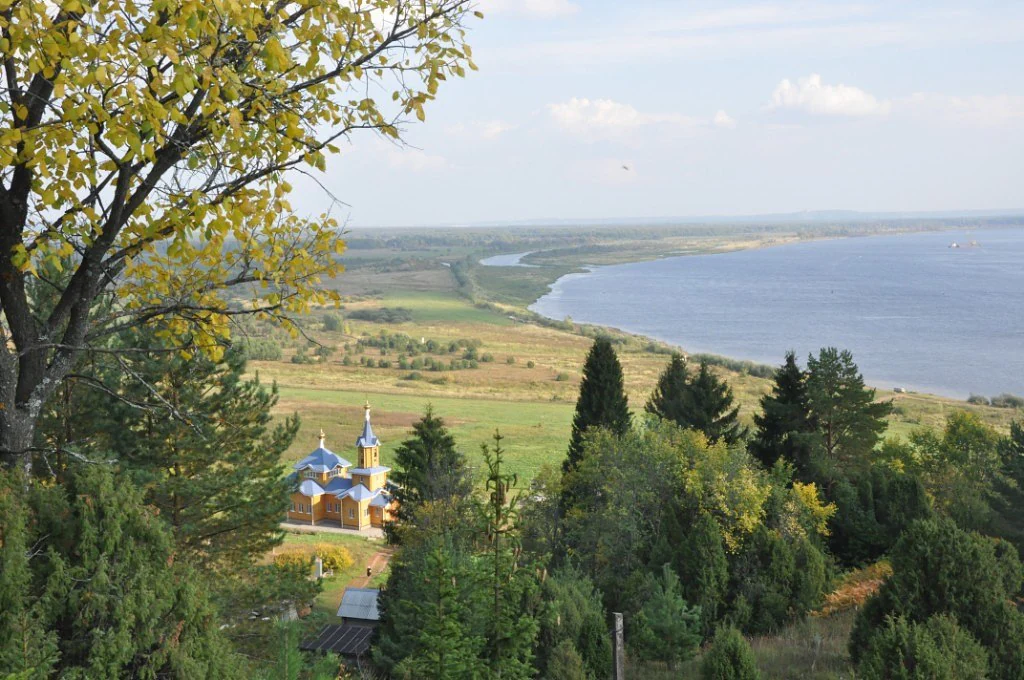 Panoramic landscape view of Nechkinsky National Park with river valley, forested hills, and a church with blue domes under a partly cloudy sky