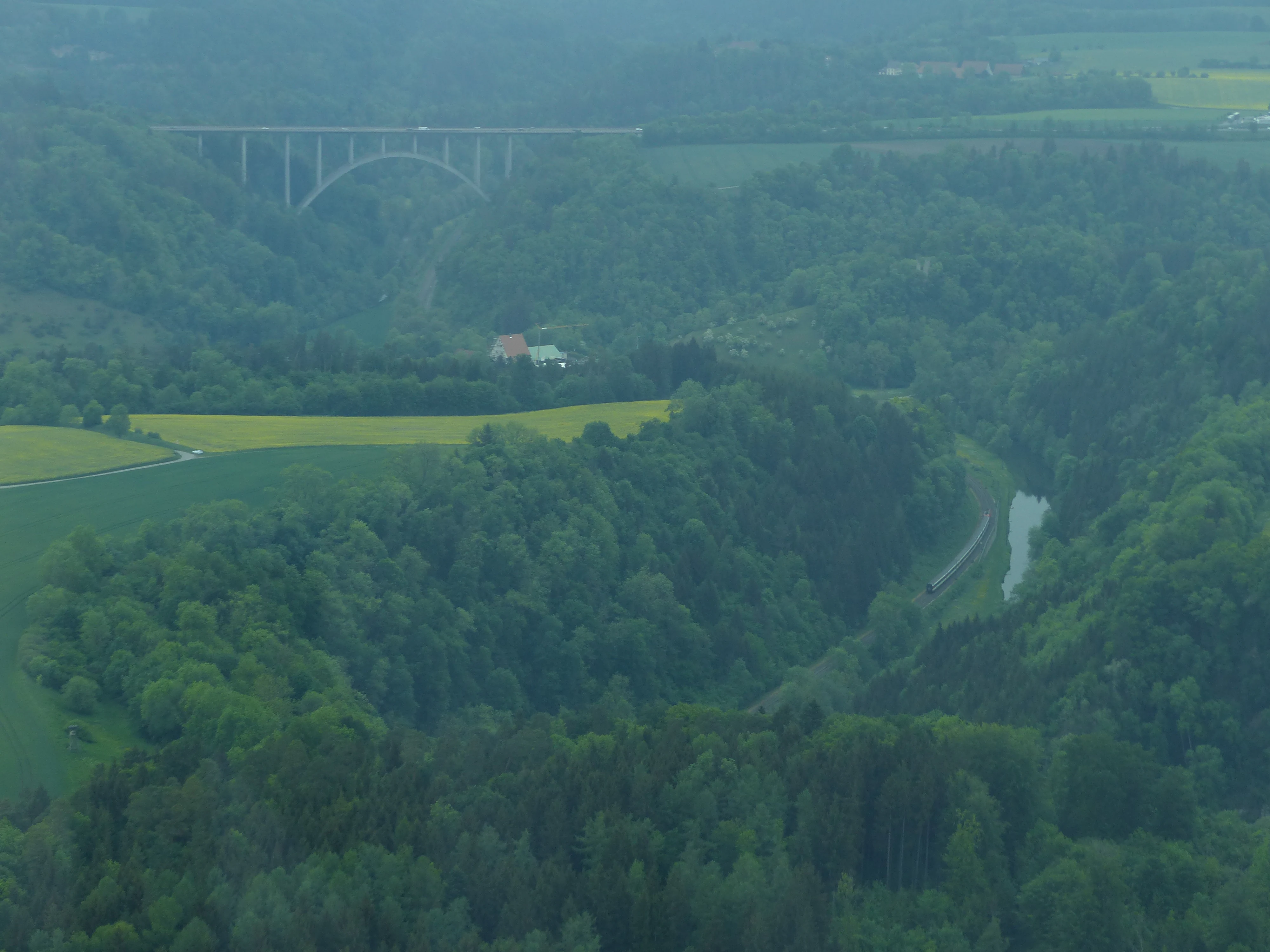 Hazy landscape view of a river valley with a large arched bridge in the background, a winding river through forested hills, a railway line with a train, and agricultural fields.