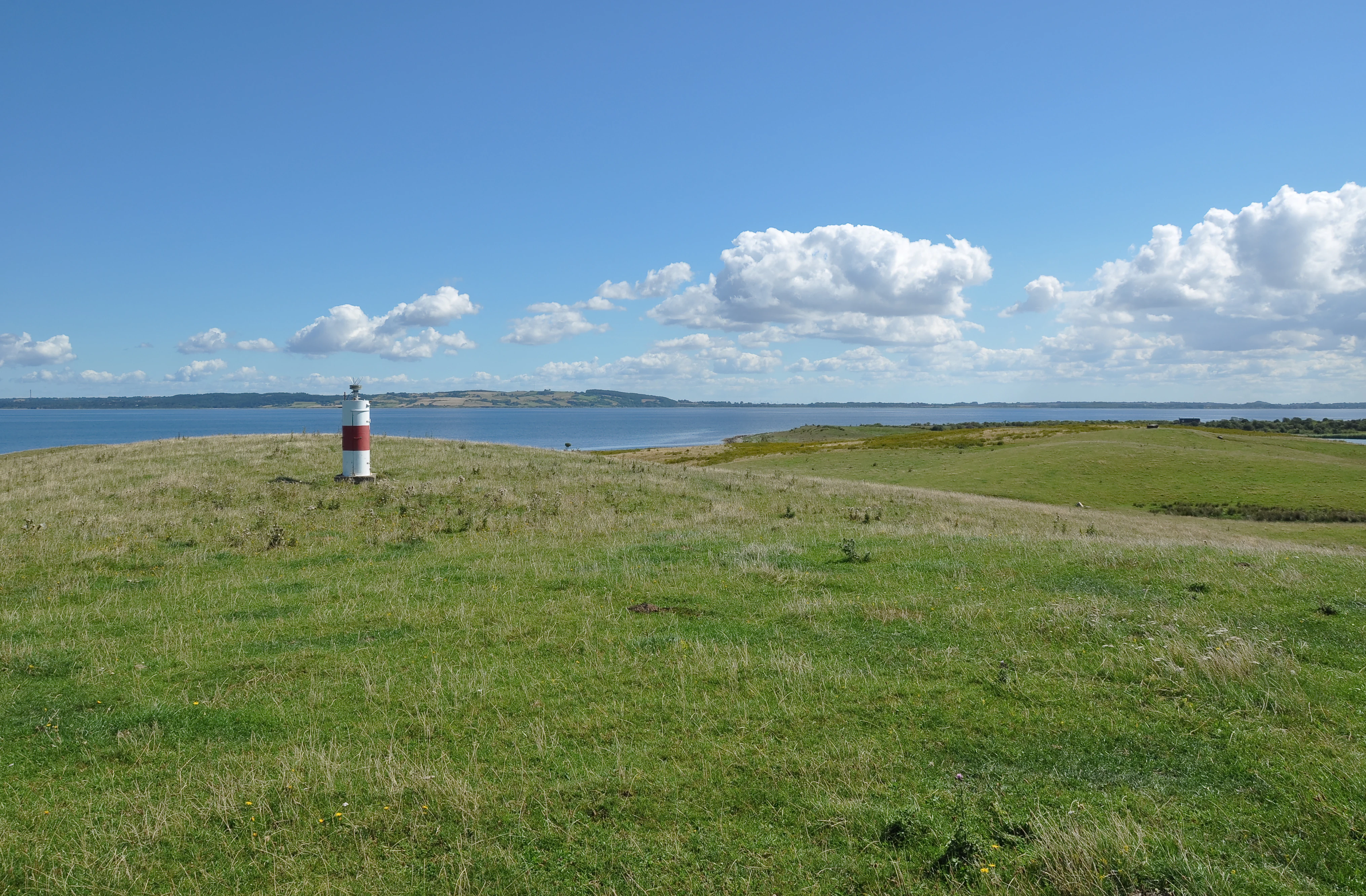 A red and white navigational beacon stands on a grassy, open hillside overlooking the sea under a clear blue sky with white clouds.