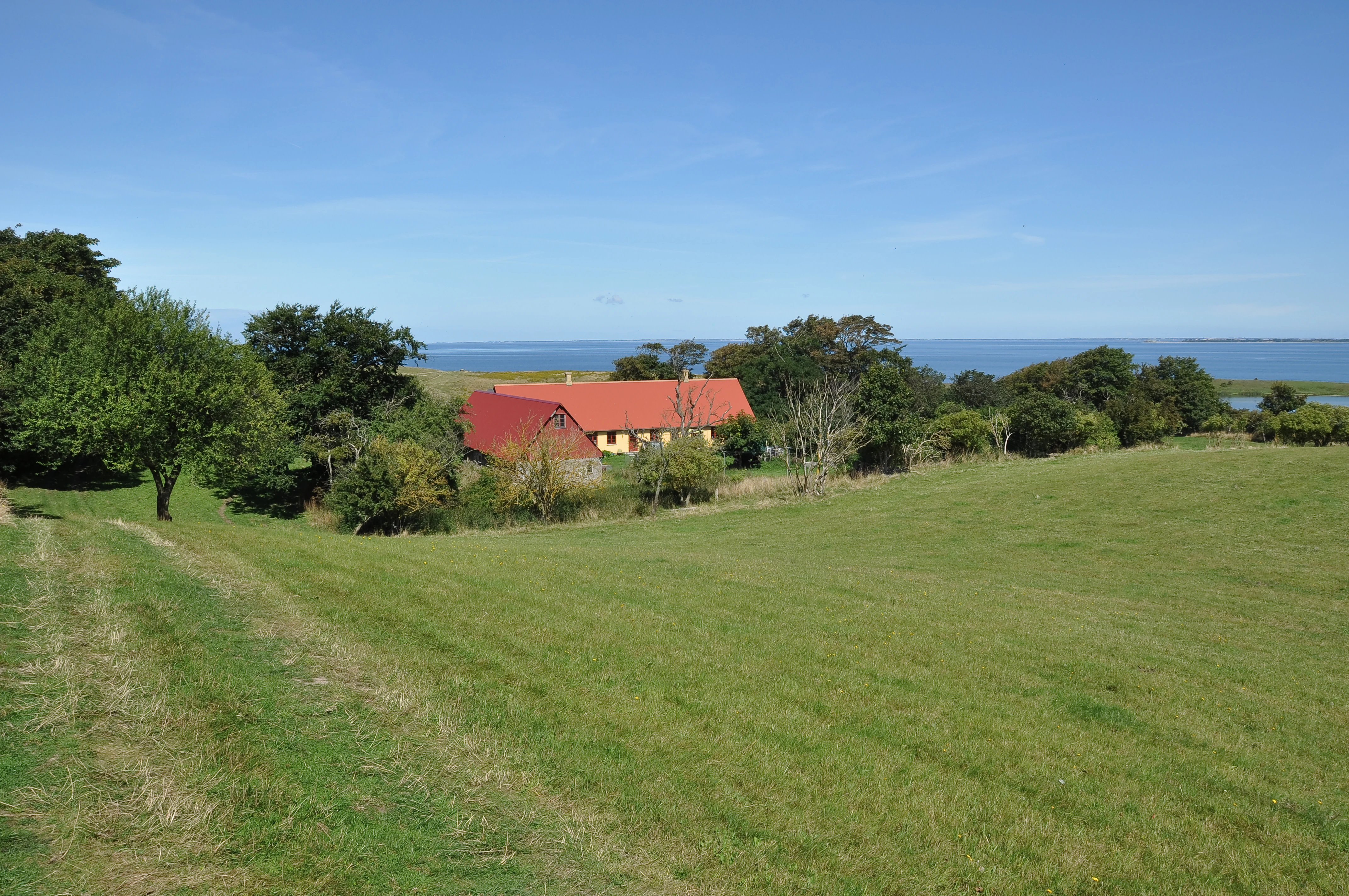 A wide view of a rural landscape on Nekselø, Denmark, featuring a traditional farmhouse with a red roof, surrounded by green fields and trees, with the sea visible in the background under a clear blue sky.