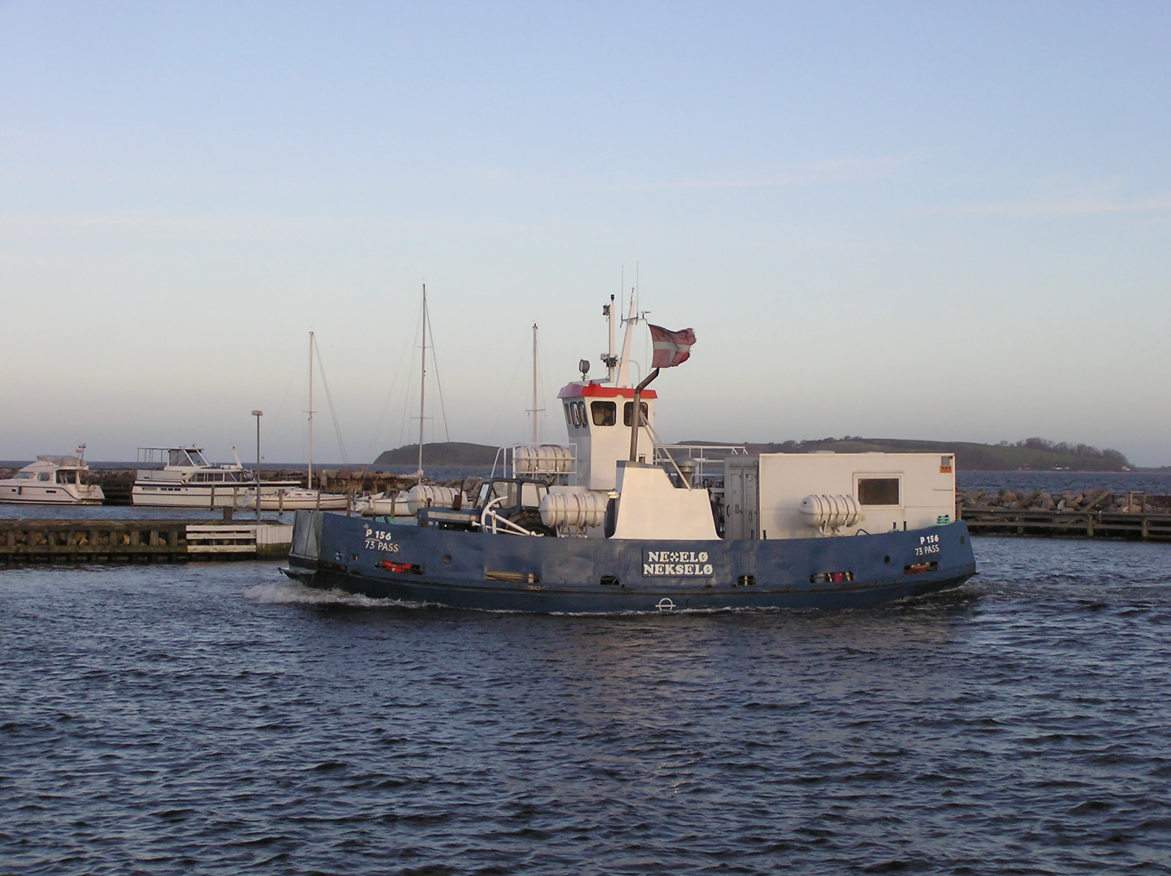 A blue ferry boat named Nekselø crossing calm water with a marina in the background.