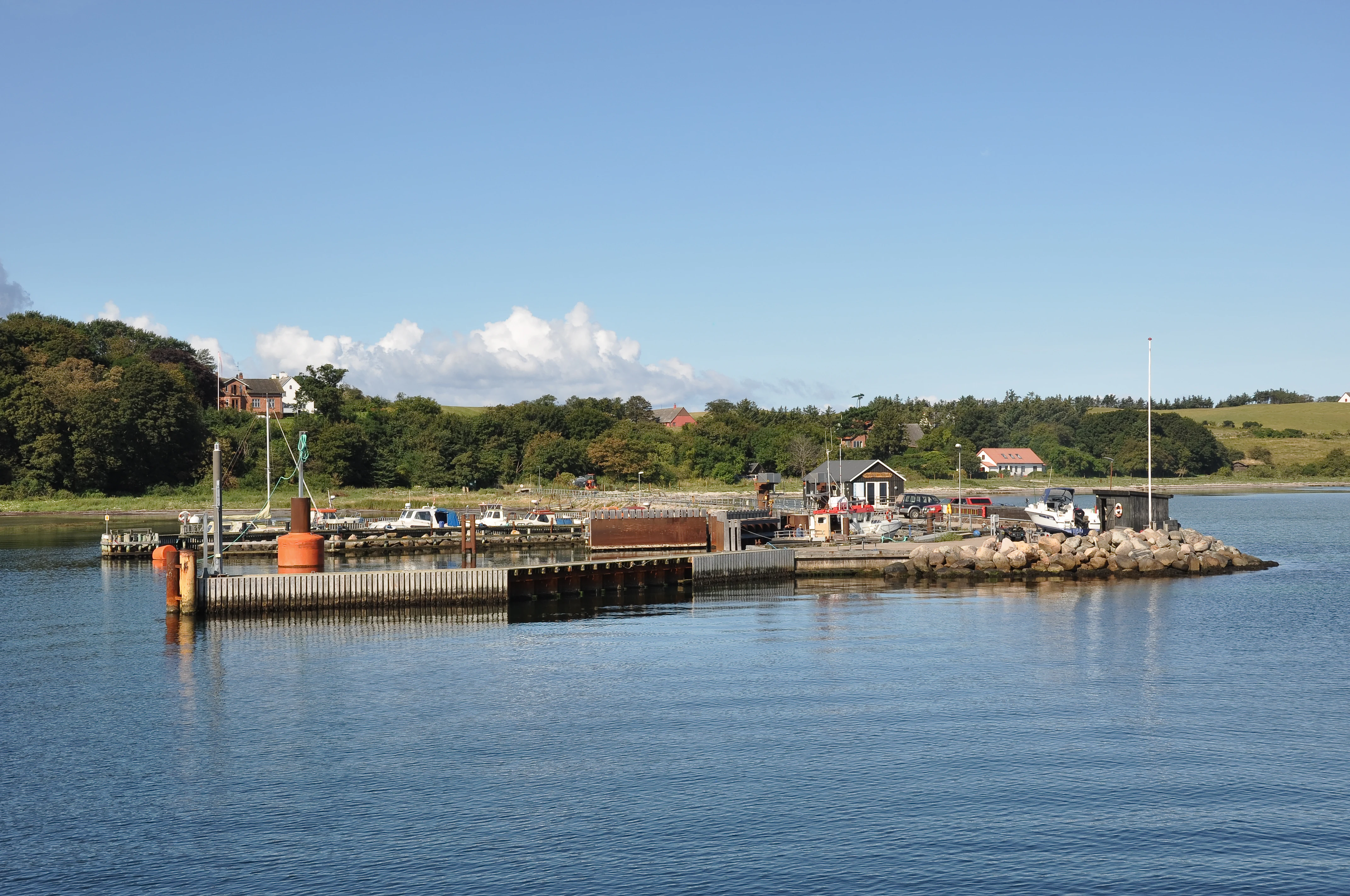 A small ferry landing with concrete piers and small buildings on the shoreline of Nekselø island, Denmark, viewed from the water.