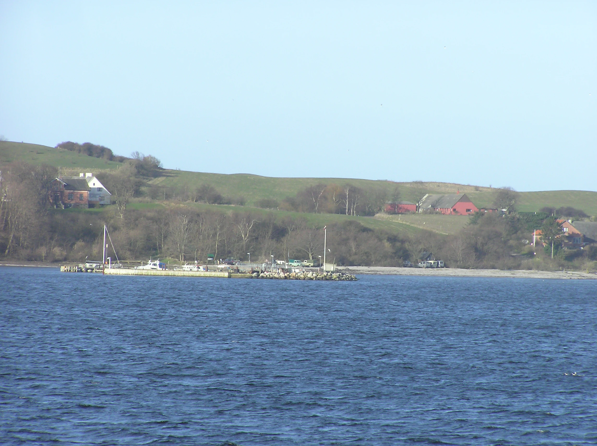A wide shot of a small harbor and concrete pier extending into calm water, with green rolling hills, a few small houses, and a red building visible on the coastal hillside in the background.