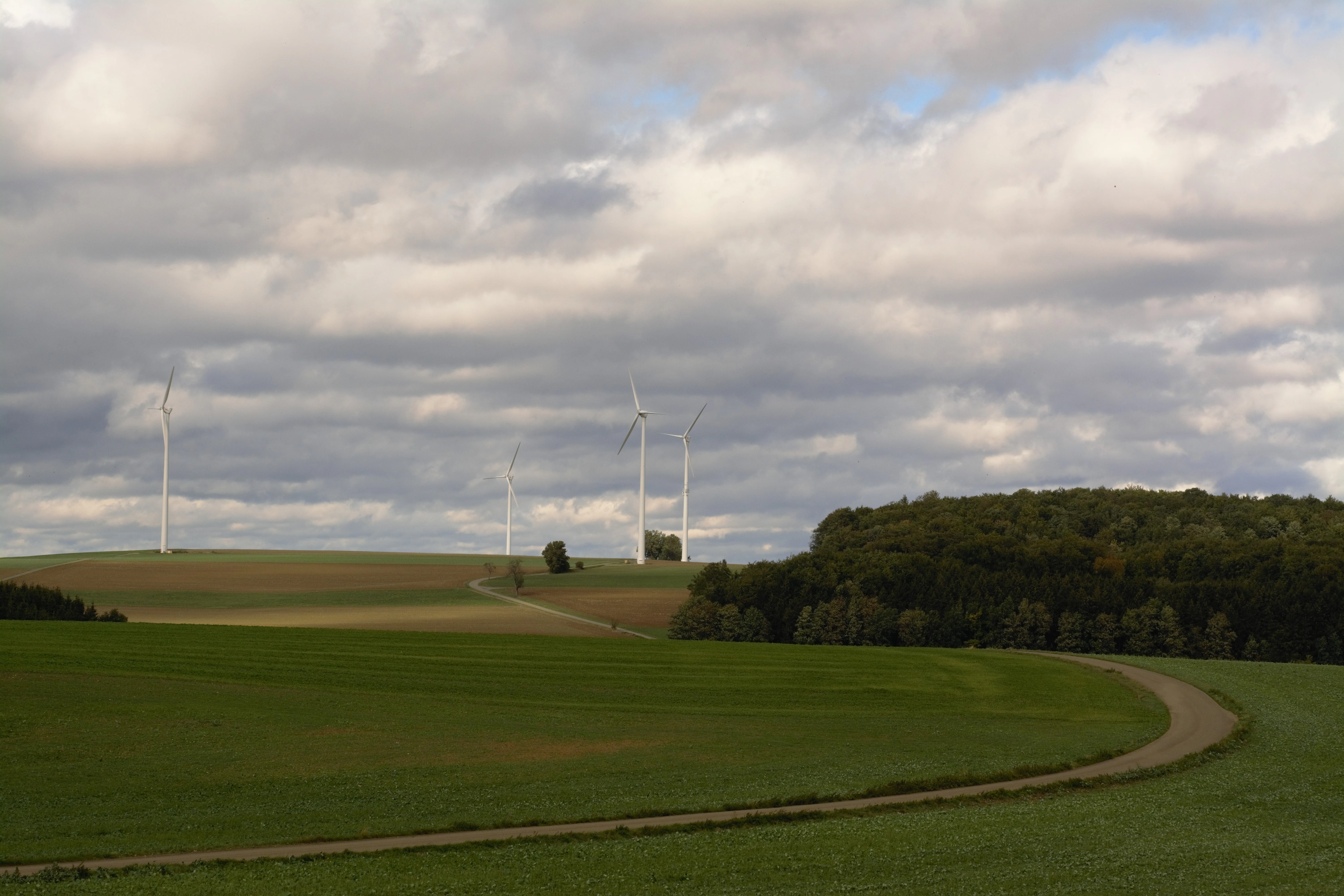 A wide view of a rolling green agricultural landscape with a dirt path winding through fields and wind turbines in the distance under a cloudy sky.