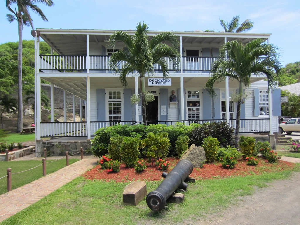 A two-story white historic building with a wrap-around balcony, identified as the Dockyard Museum, with a cannon displayed in the foreground garden.