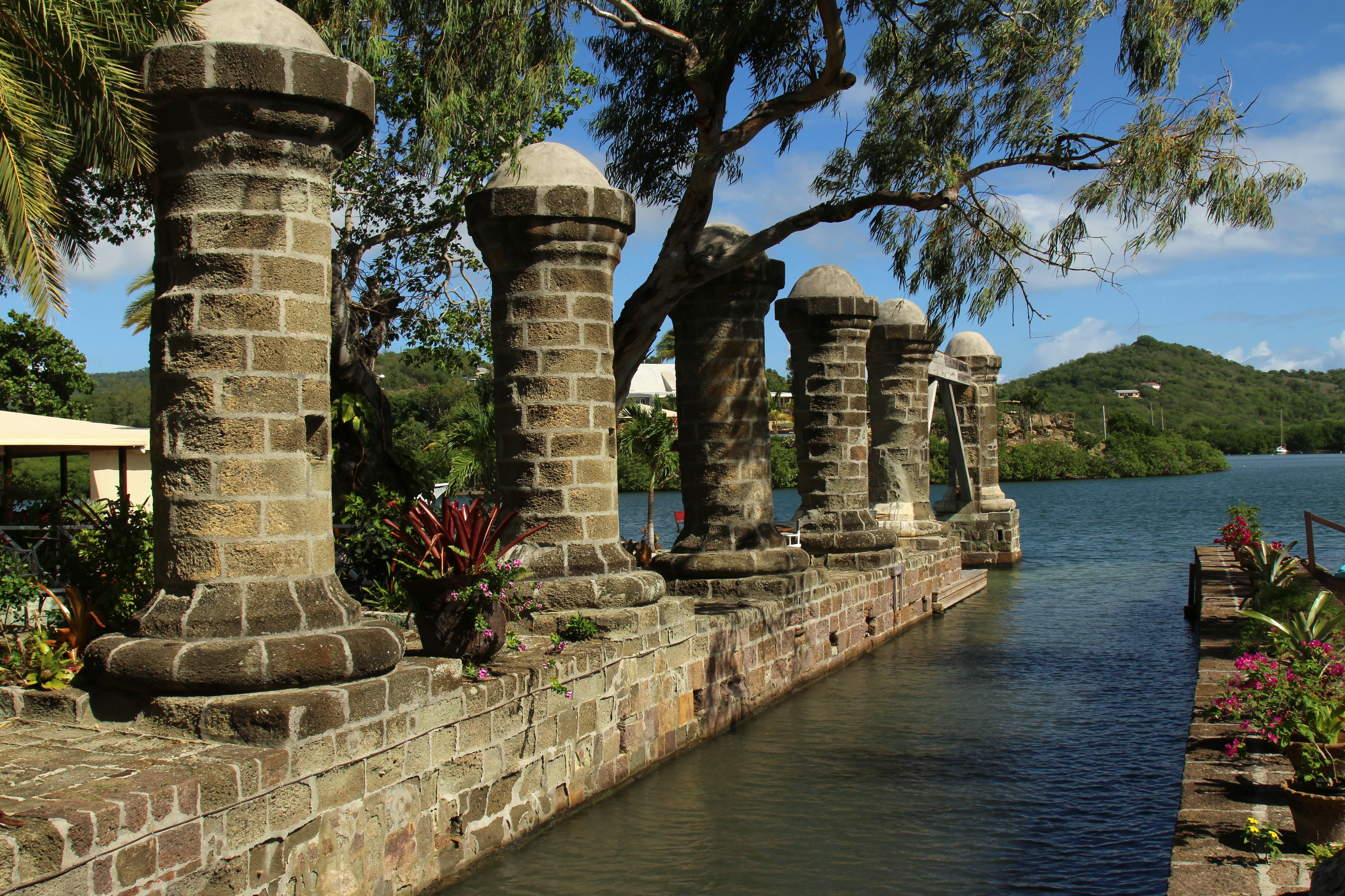A row of stone pillars stands along a harbor wall at Nelson's Dockyard, with blue water and green hills visible in the background.