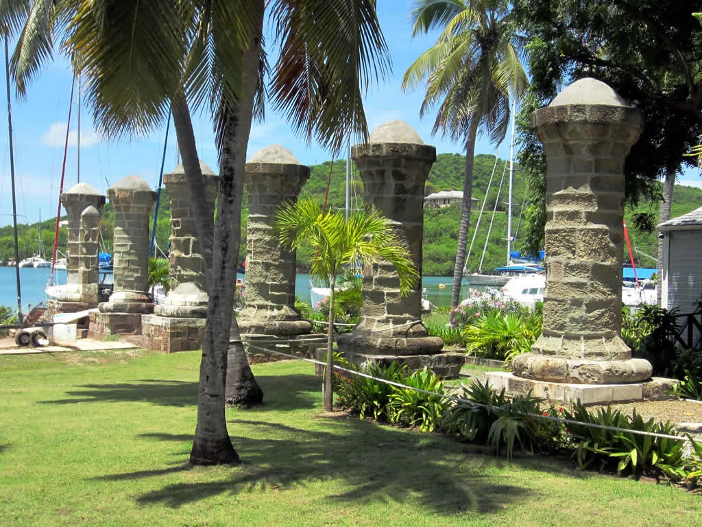 A row of several tall, weathered stone columns with rounded caps standing in a grassy area at Nelson's Dockyard in Antigua, with palm trees and sailboats visible in the background.