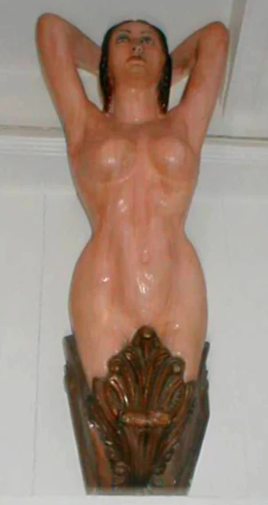 A close up vertical shot of a wooden ship figurehead depicting a female torso with arms raised, mounted on a wall against a plain white background.