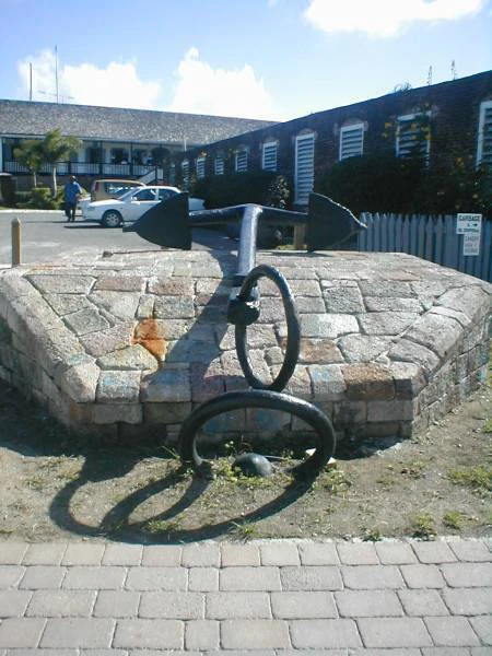 A large, black, antique metal ship anchor displayed on a low, raised stone platform in an outdoor courtyard.