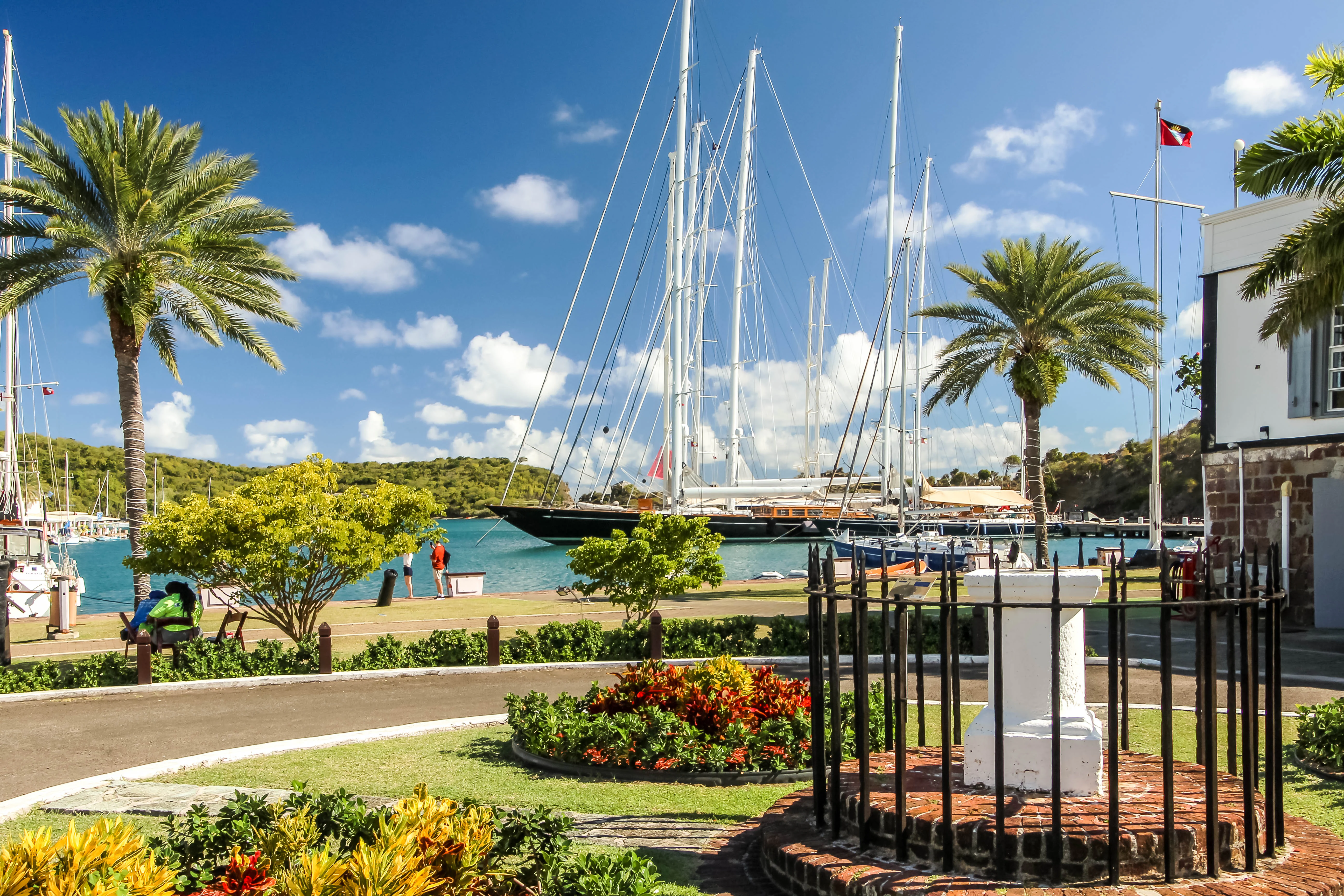 A sunny view of English Harbour with a large sailing yacht docked, palm trees, and historic stone architecture in the foreground.