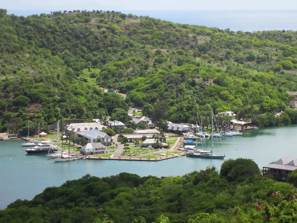 A high-angle view of a historic maritime facility with colonial-style buildings along a harbor waterfront, surrounded by lush green hills and anchored yachts.