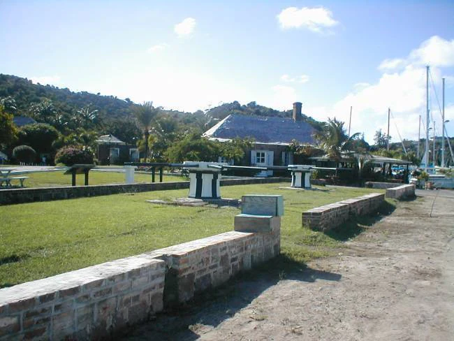 A wide view of the grassy area at Nelson's Dockyard, featuring low stone walls, white painted mooring bollards, and 18th-century stone buildings in the background.