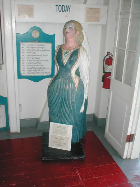 A painted wooden figurehead of a woman in a long teal dress stands on display in a museum room, with an information sign in front of her.