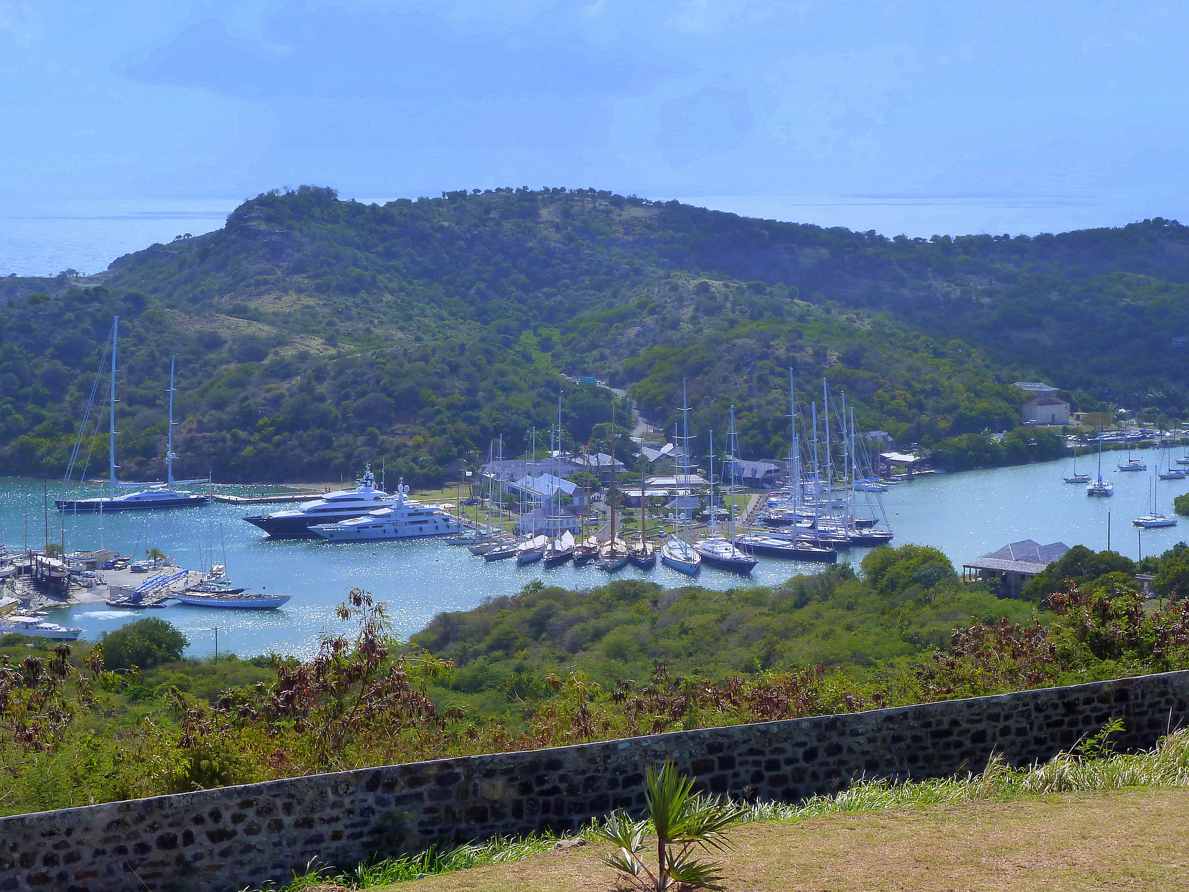 A wide angle view from a stone-walled vantage point looking down at a harbor filled with yachts and sailboats, surrounded by lush green hills.