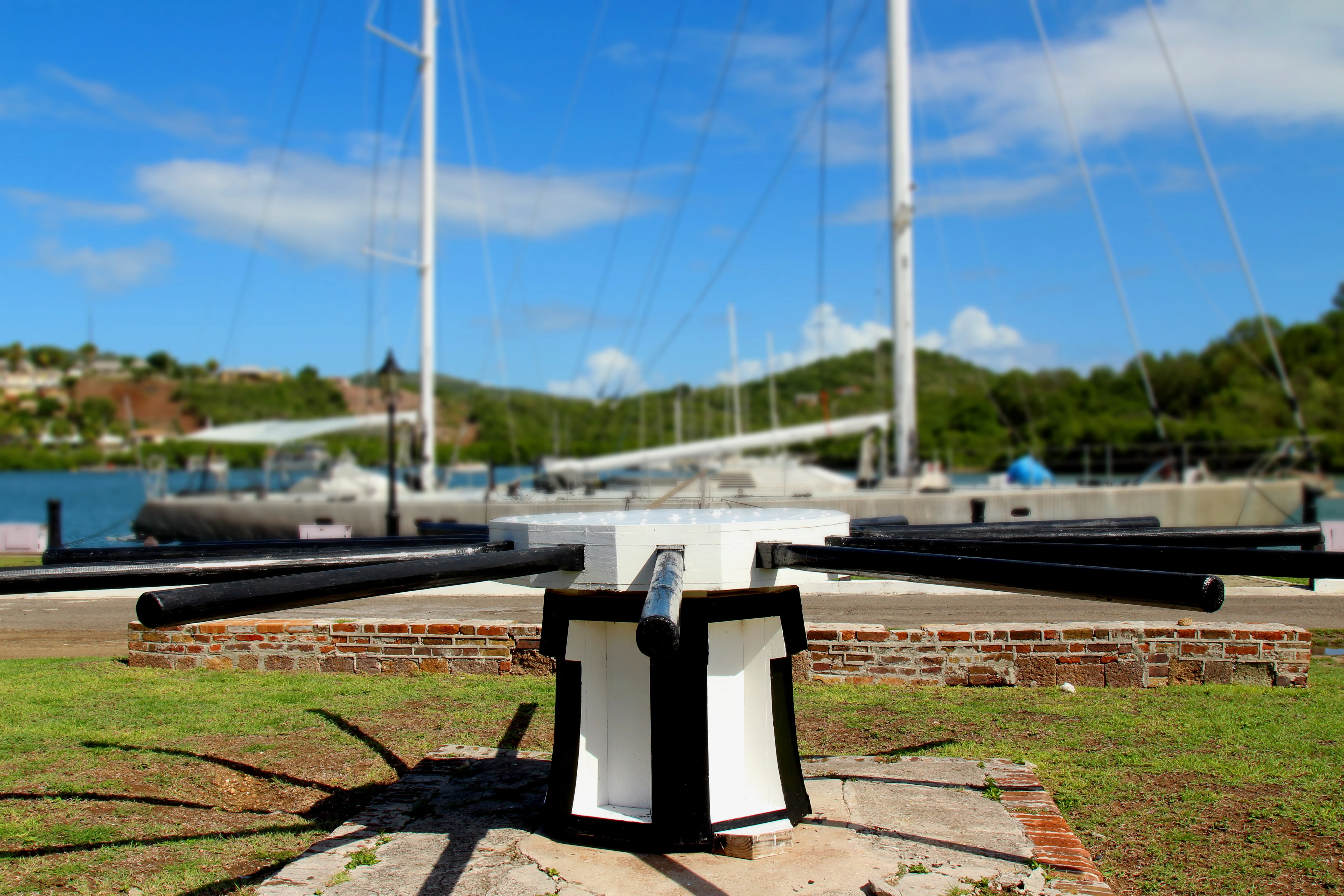 A white and black historic mechanical capstan stands in the foreground, with a large sailing vessel docked in the background at English Harbour.