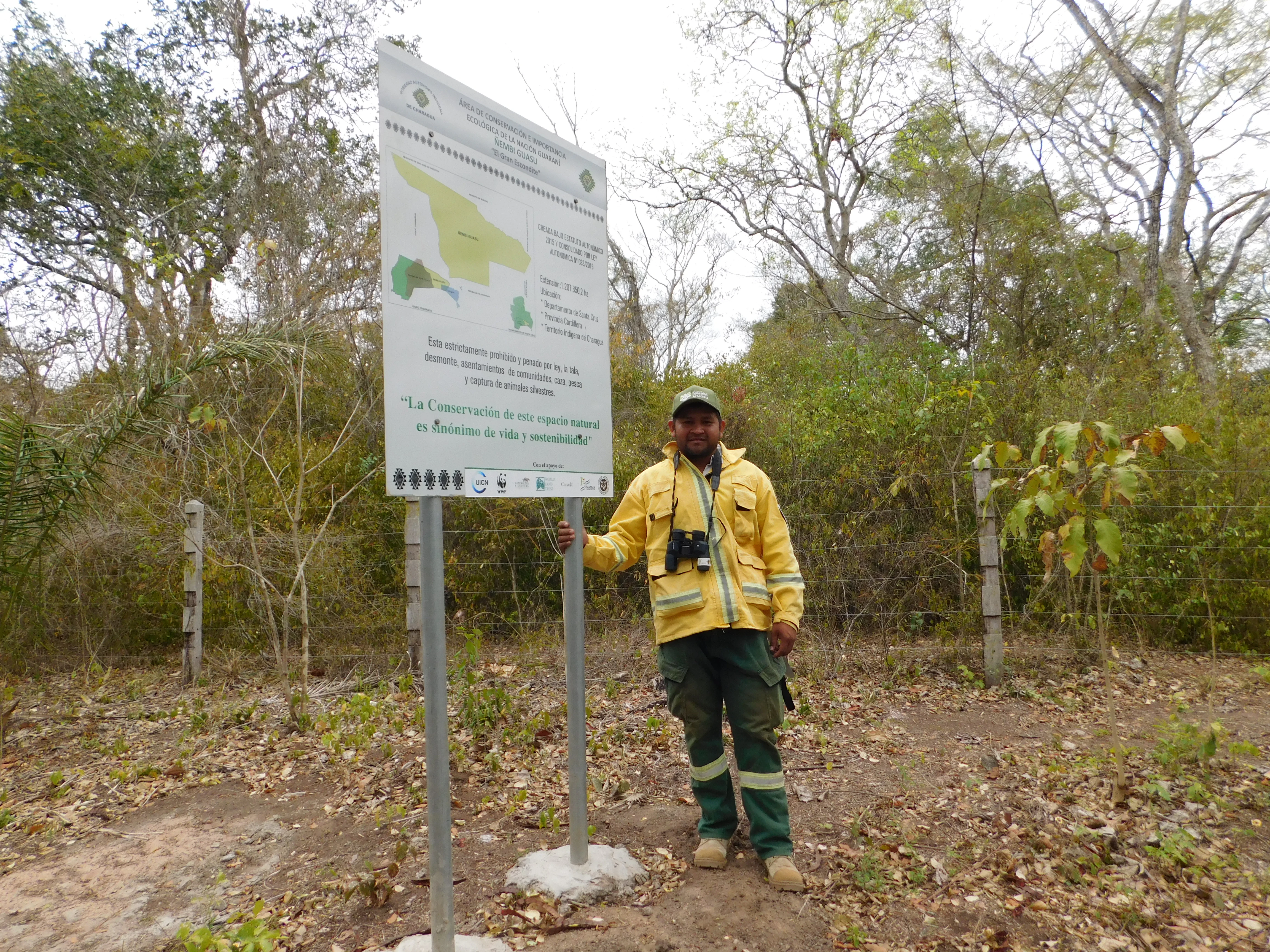 A person in a yellow uniform and dark pants standing next to a tall white informational sign in a dry forest setting.