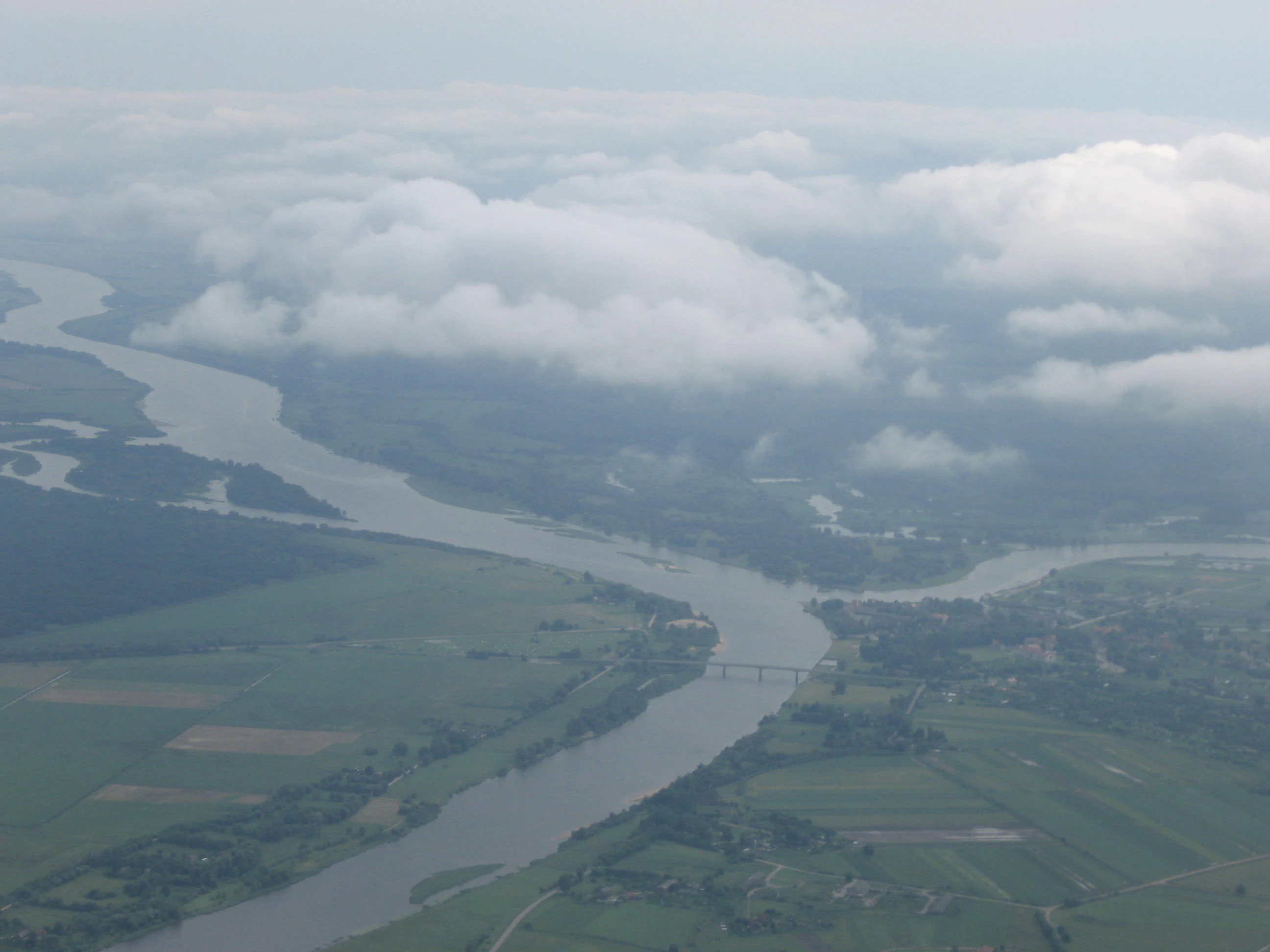 An aerial shot of a wide, winding river flowing through a flat, green landscape with distant patches of trees, partially obscured by low-hanging clouds.