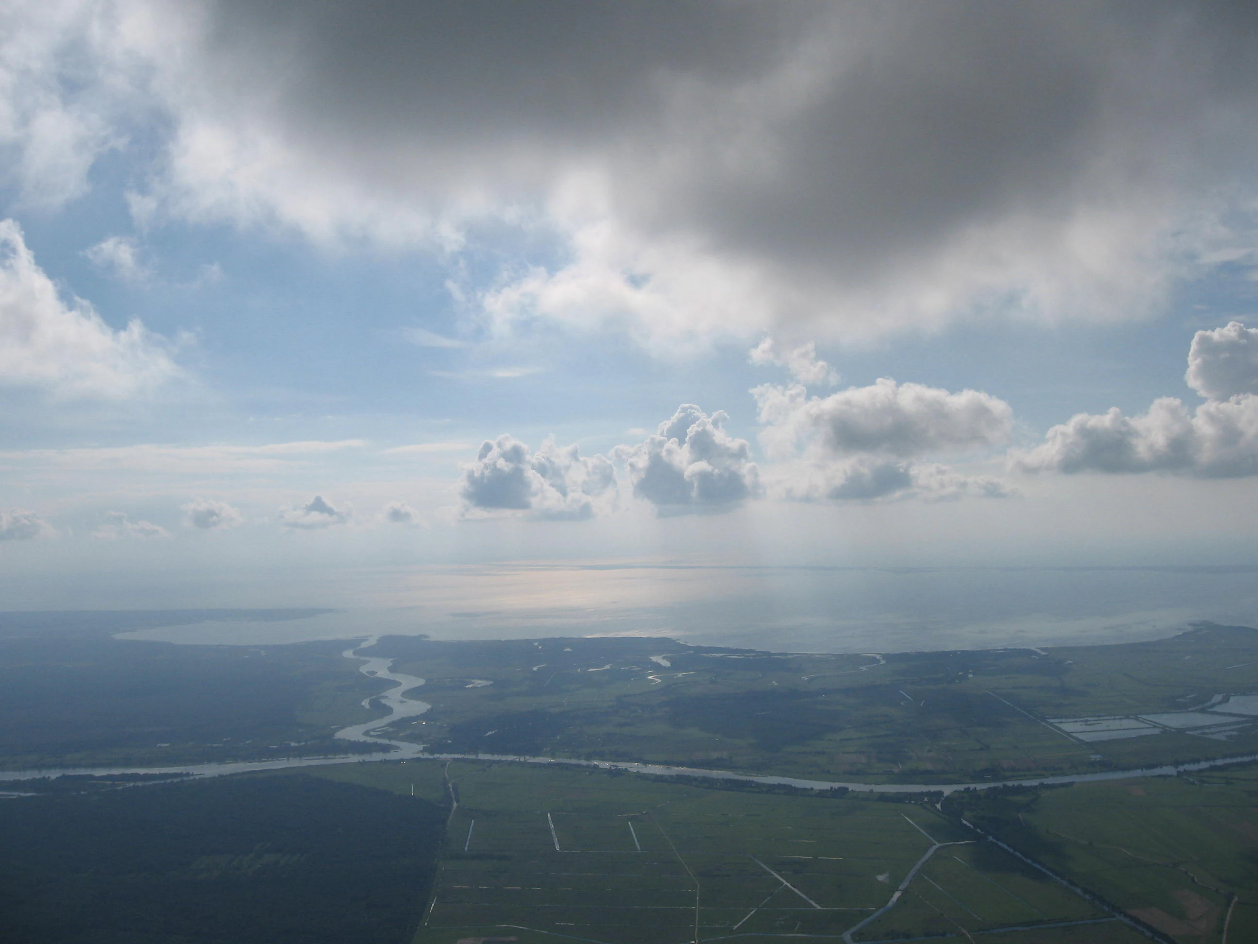 Aerial view looking over flat, green wetlands and river channels as they flow into a large, bright body of water under a cloudy sky.
