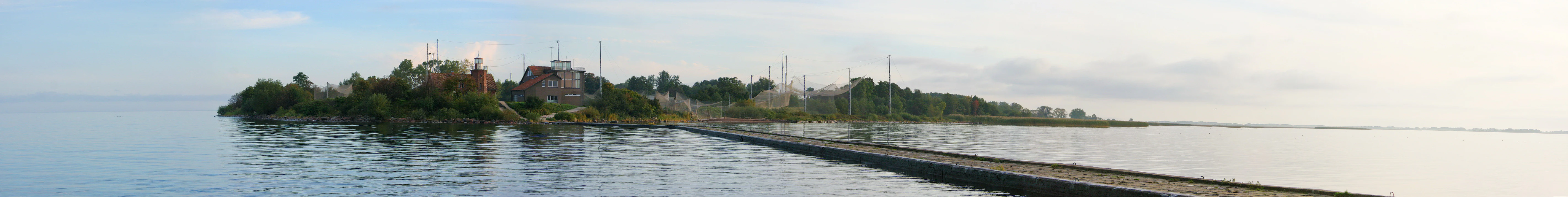 A wide panoramic view showing a low-lying, tree-covered cape extending into calm water, with a long pier or jetty stretching out from the shore toward the horizon.