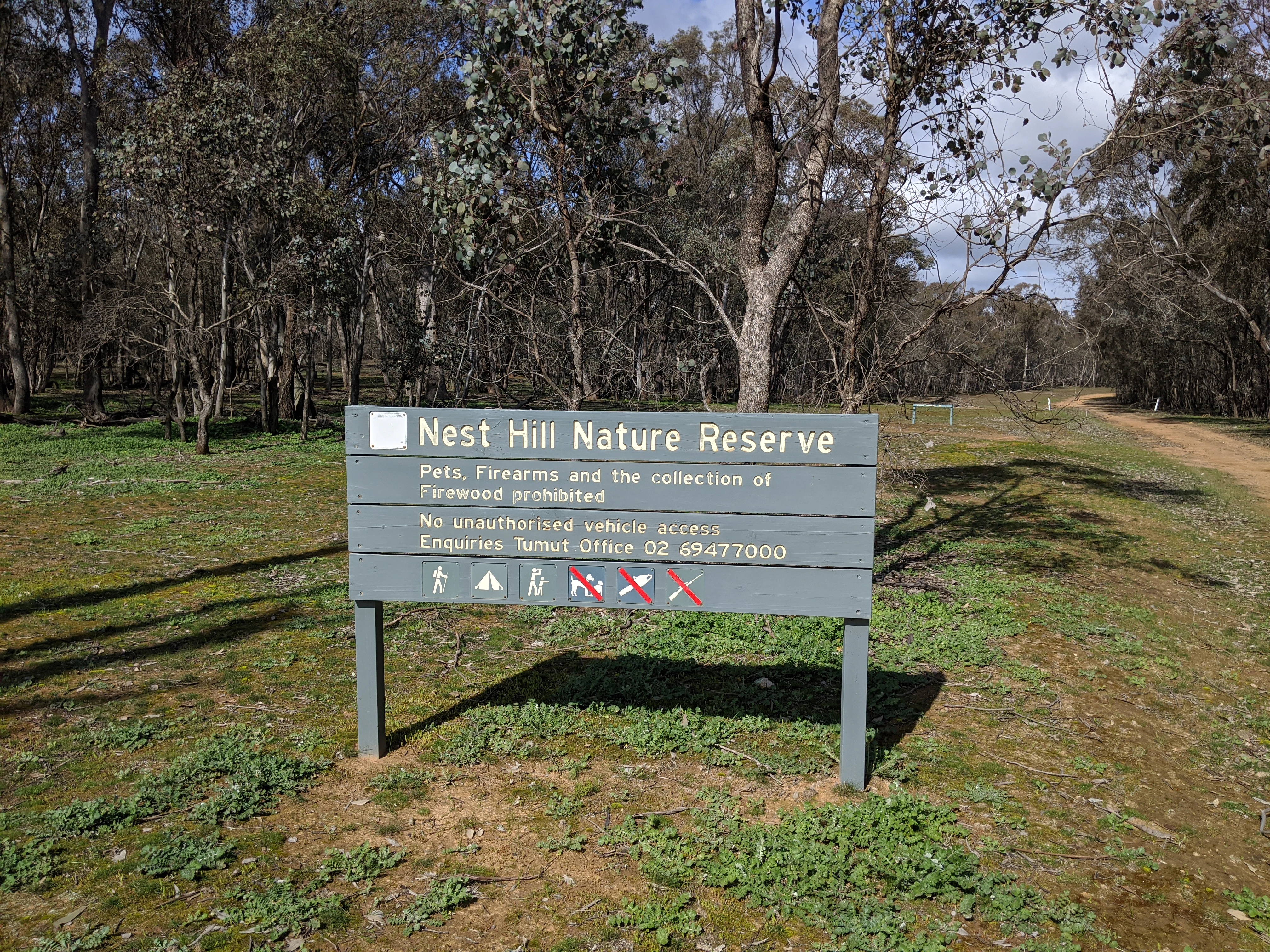 A grey park entrance sign for Nest Hill Nature Reserve standing in a dry, sparse woodland area.